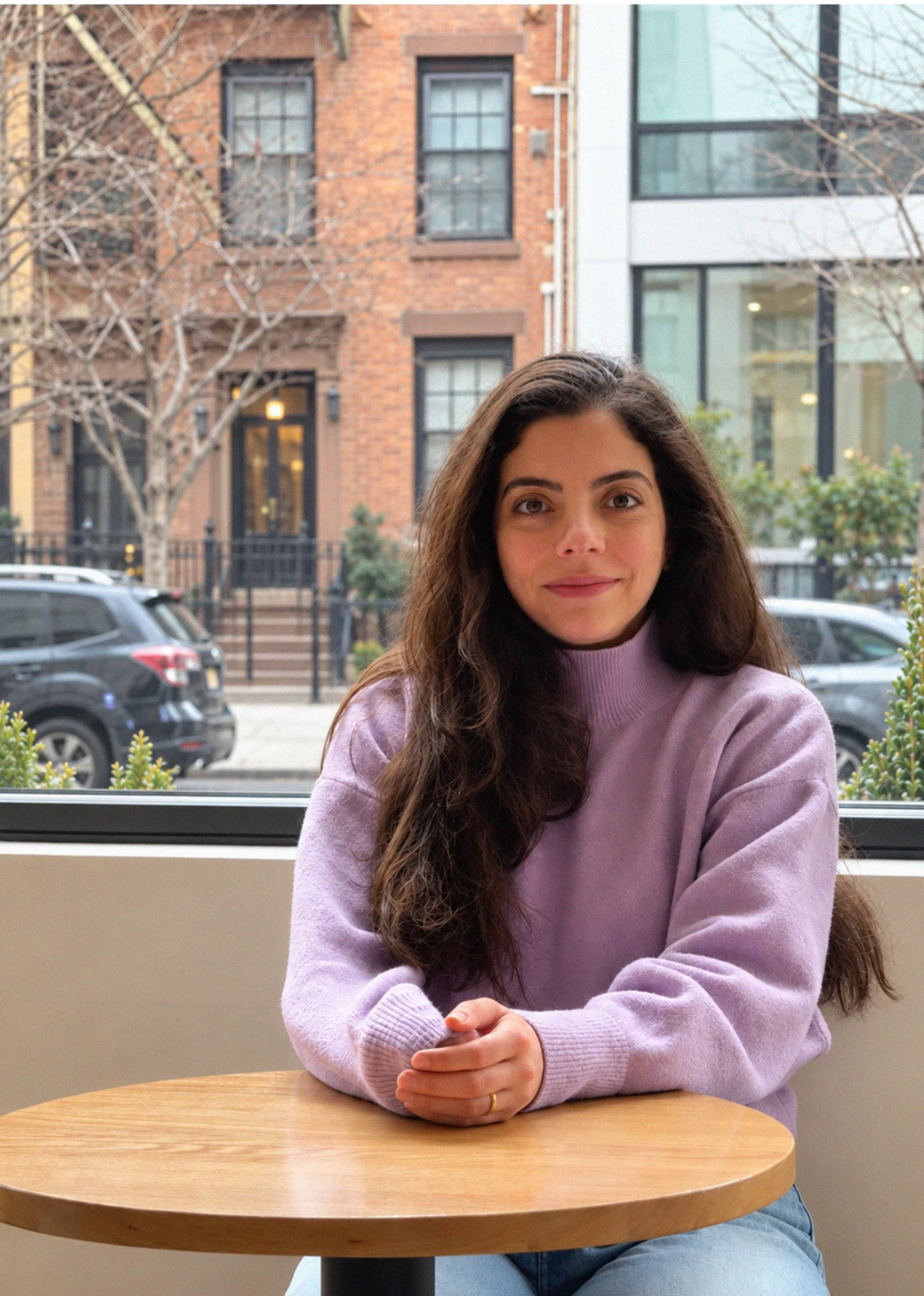 A woman with long brown hair wearing a lavender sweater sitting at a round wooden table in a cafe. She is smiling gently and has her hands folded on the table. Behind her is a large window showing a street scene with parked cars, leafless trees, and modern buildings.