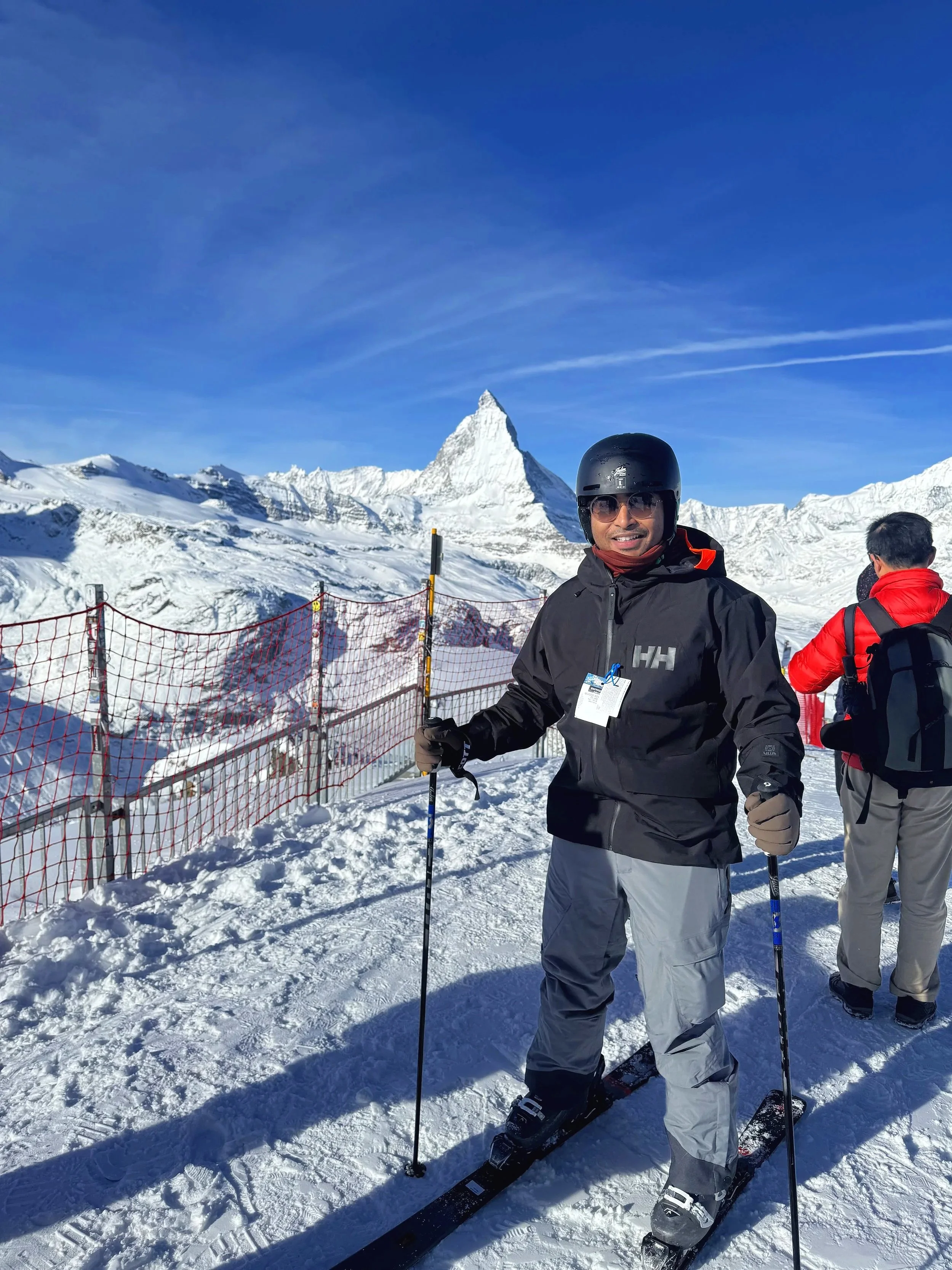 Sreyan ski gear holding ski poles standing on snow, with snow-covered mountains and the Matterhorn peak in the background, under a clear blue sky.