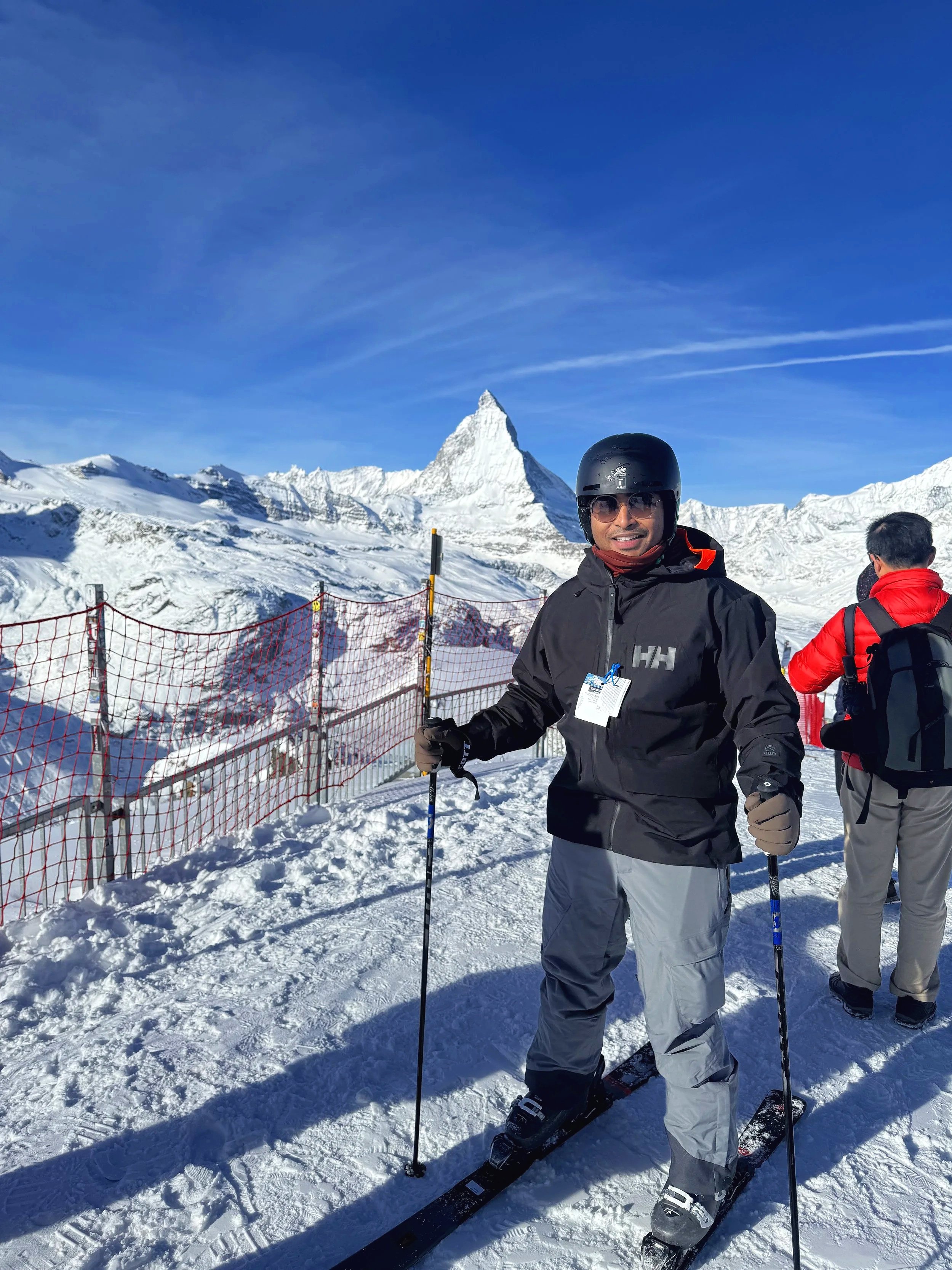 Sreyan ski gear holding ski poles standing on snow, with snow-covered mountains and the Matterhorn peak in the background, under a clear blue sky.