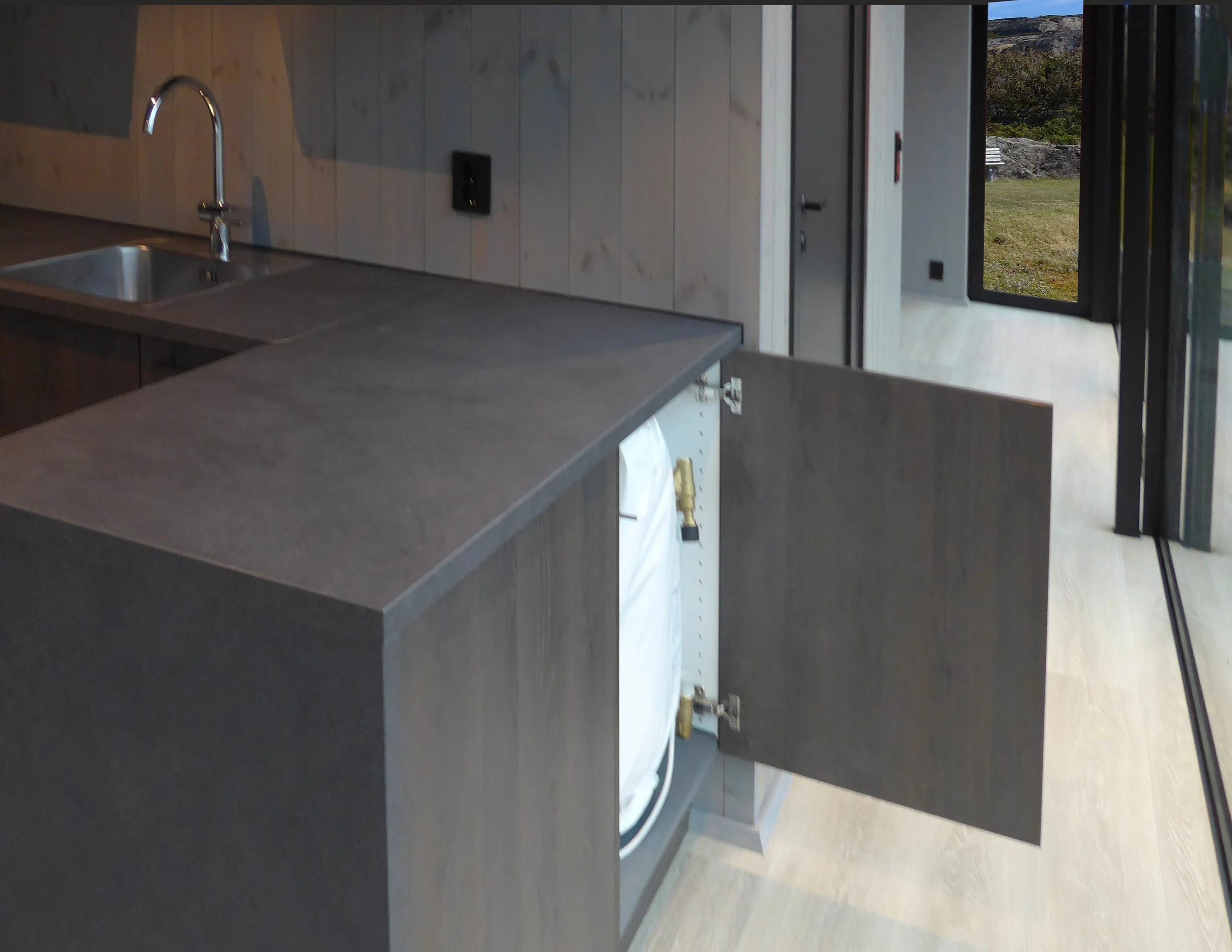 Indoor scene showing a kitchen with a countertop, sink with a faucet, and wood-paneled wall. There is a door or window with outdoors visible, and some objects and fixtures near the bottom of the image.
