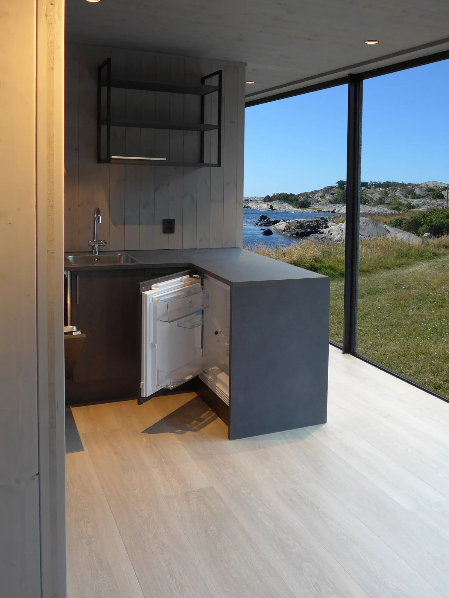 Interior view of a modern kitchen with a small sink, open shelving, and a view of a rocky shoreline and blue sky through large glass windows.