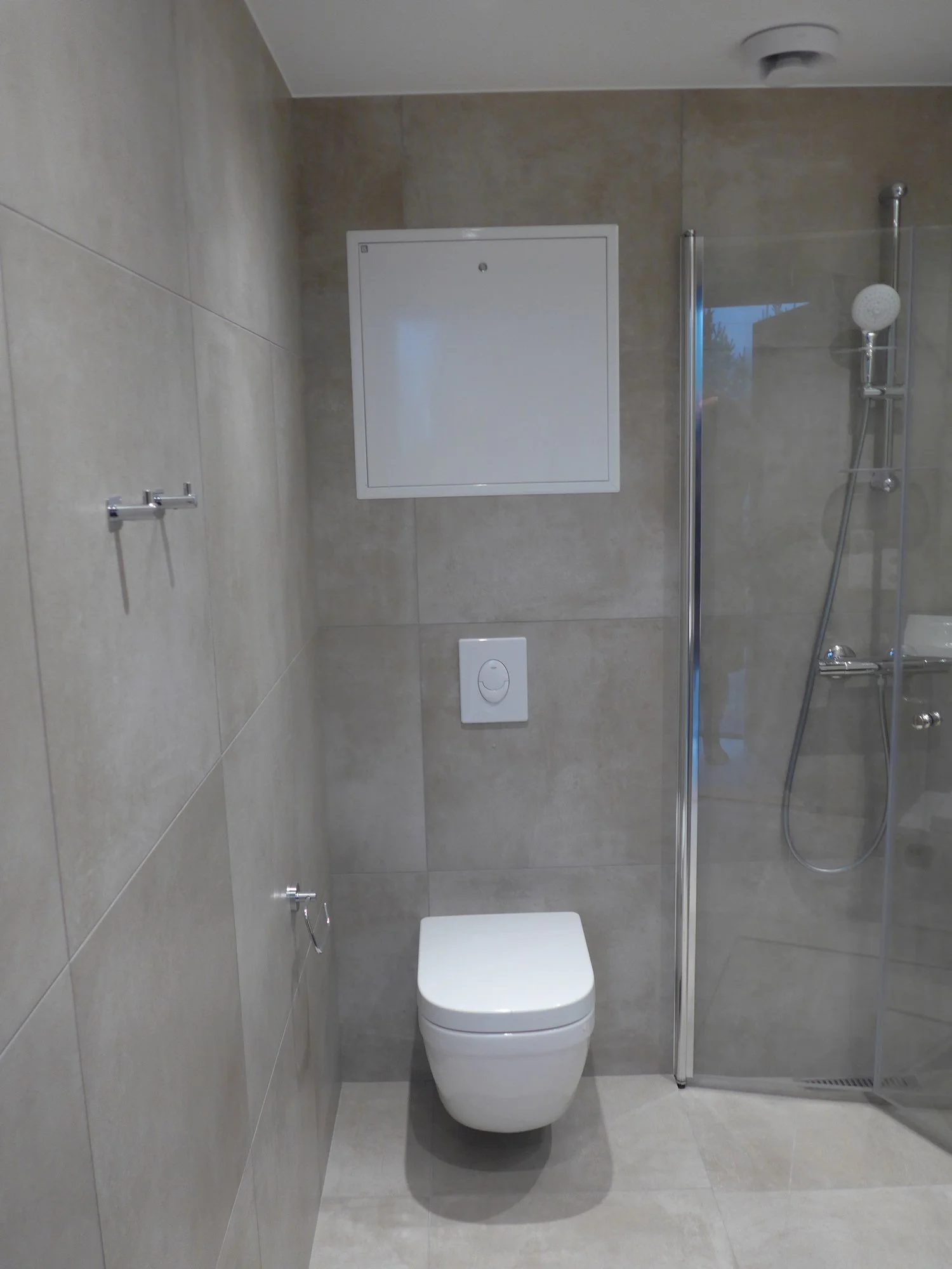 Modern bathroom with beige tiled walls, a wall-mounted white toilet, a small white flush panel, a glass shower door, and a shower with a handheld showerhead.