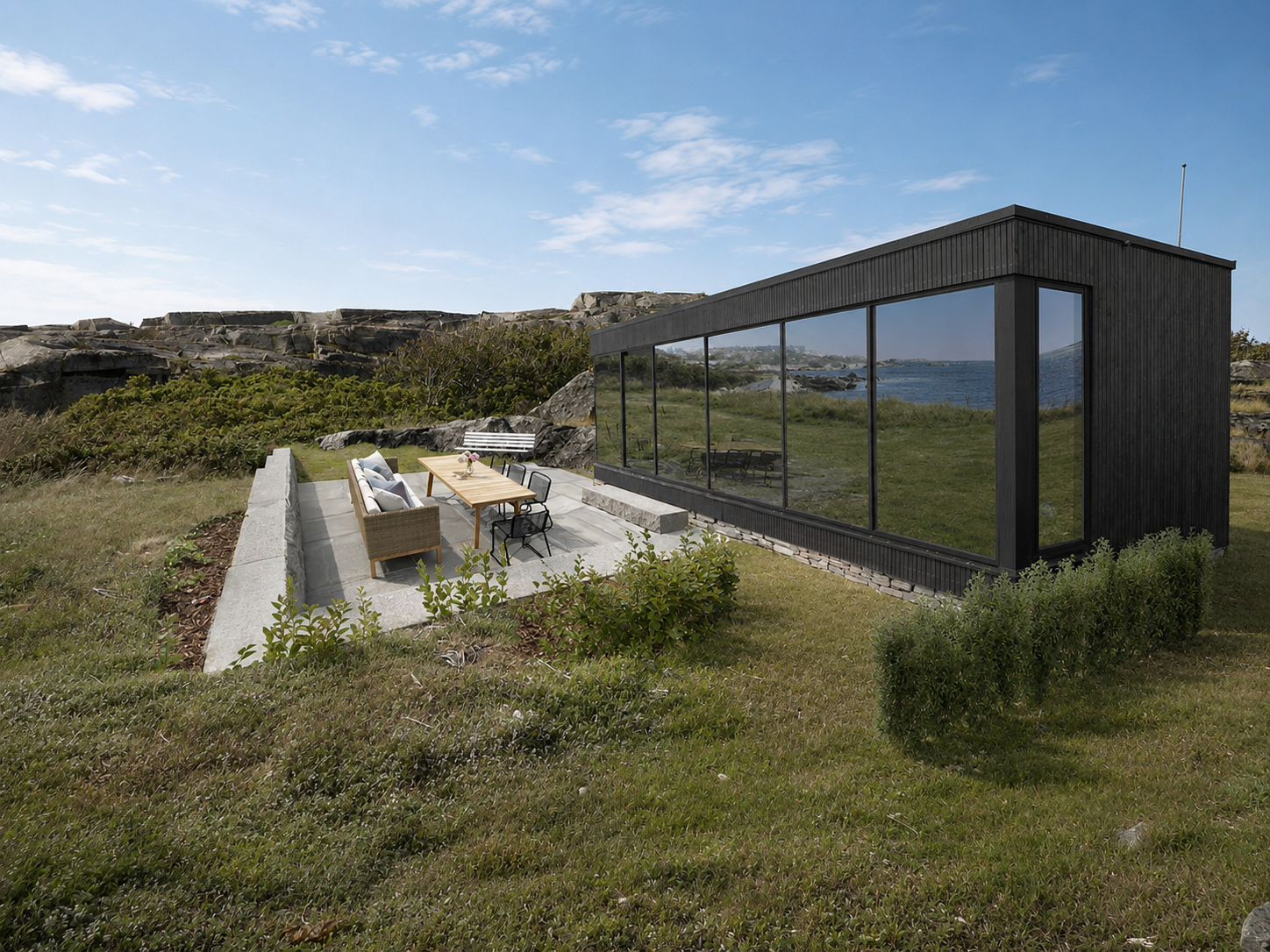 Modern black house with glass walls overlooking a grassy landscape and water, with outdoor seating and dining area on a stone patio.