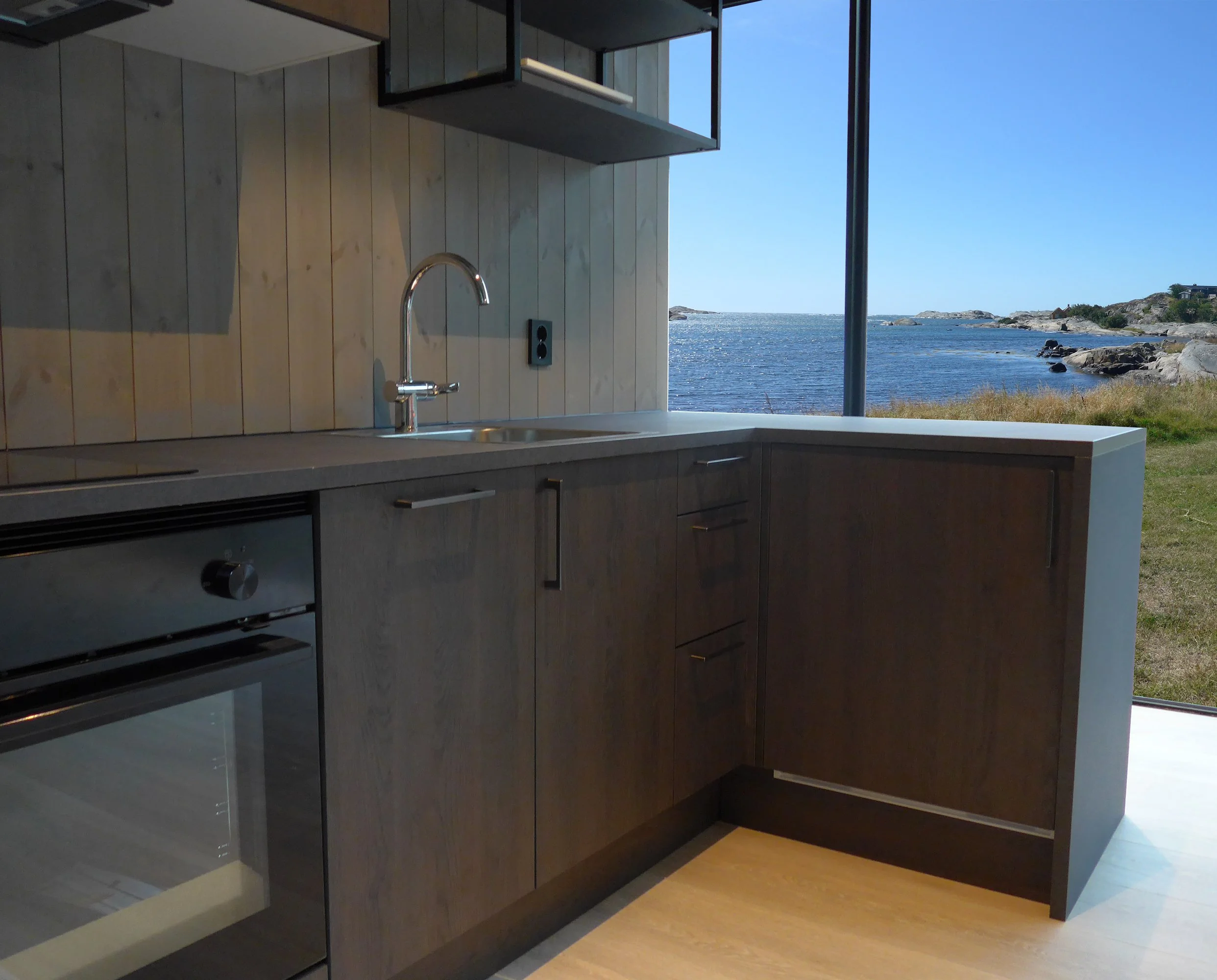Modern kitchen with dark cabinets, a light-colored countertop, a stainless steel faucet, and a window overlooking a landscape with water, rocks, and a clear blue sky.