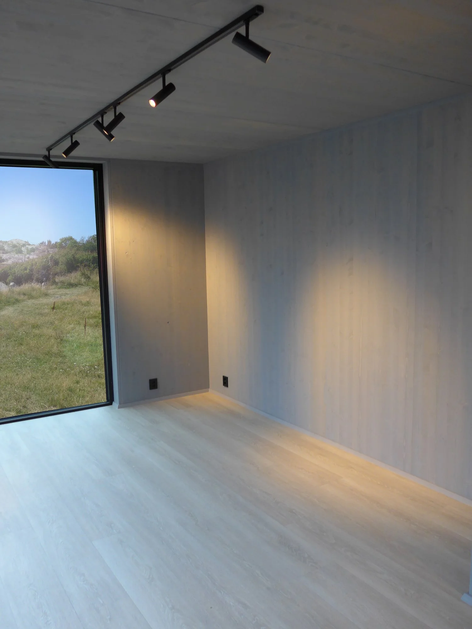 Empty modern room with light wooden flooring and textured light gray walls, featuring large floor-to-ceiling window and black ceiling track lighting with adjustable spotlights.