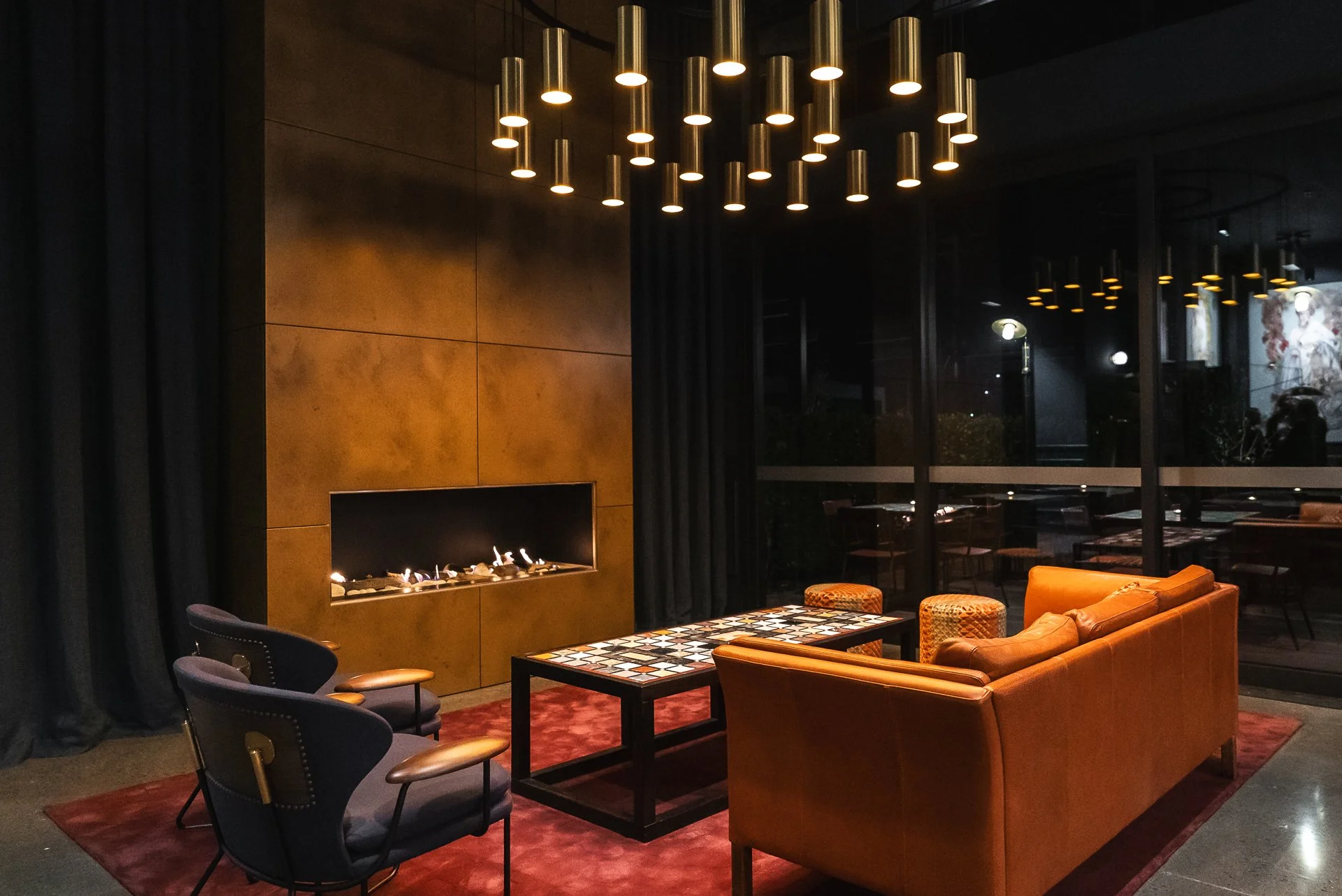 Interior of a modern living room with a fireplace, black curtains, a glass-topped coffee table, an orange sofa, and matching armchairs. Modern cylindrical pendant lights hang from the ceiling, and large windows reveal a dark outdoor scene with visible reflections.