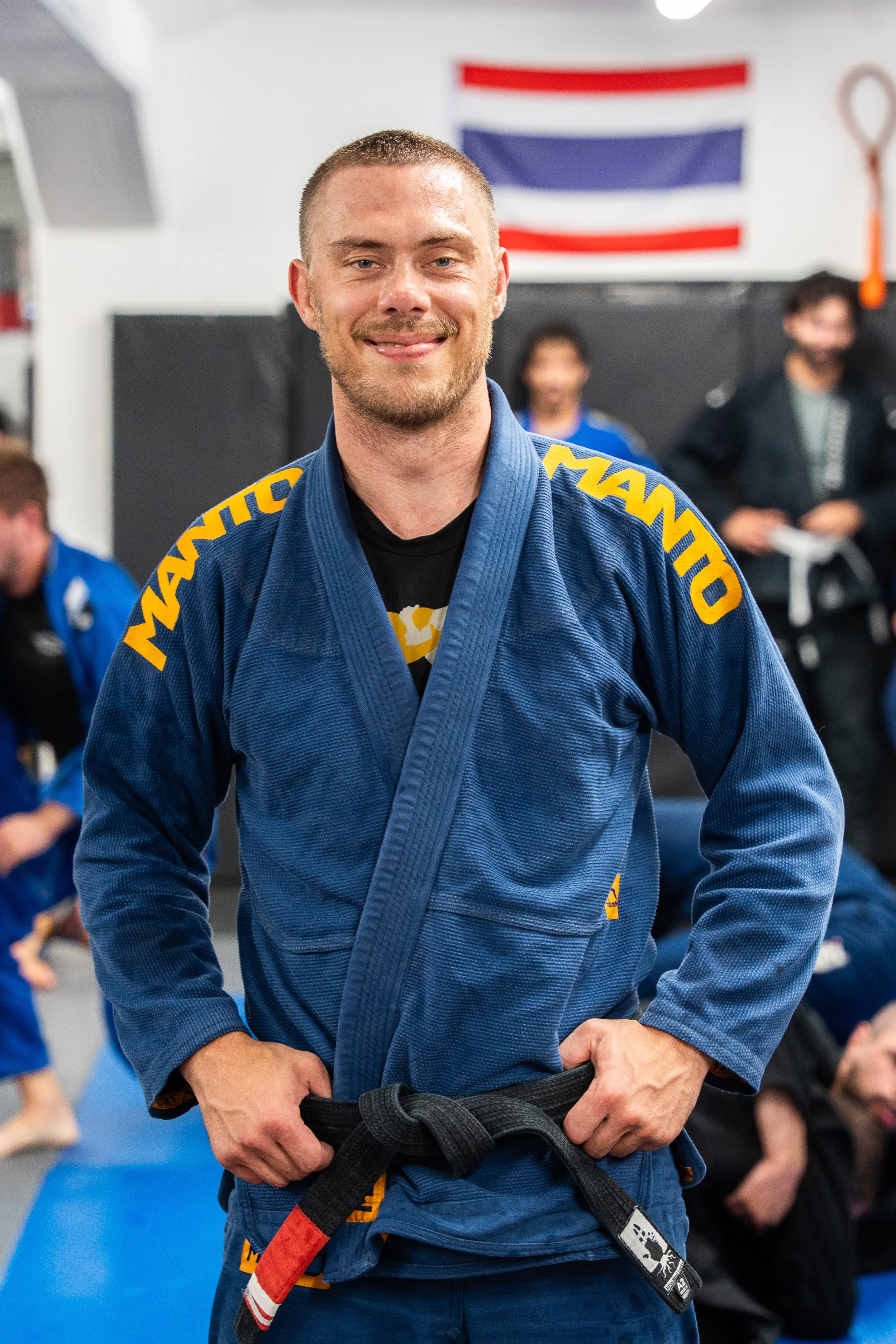 Man i blå karate gi med gul text, står i trädning på en dojo, bugar med händerna på midjan. I bakgrunden ett norskt flagga på väggen.