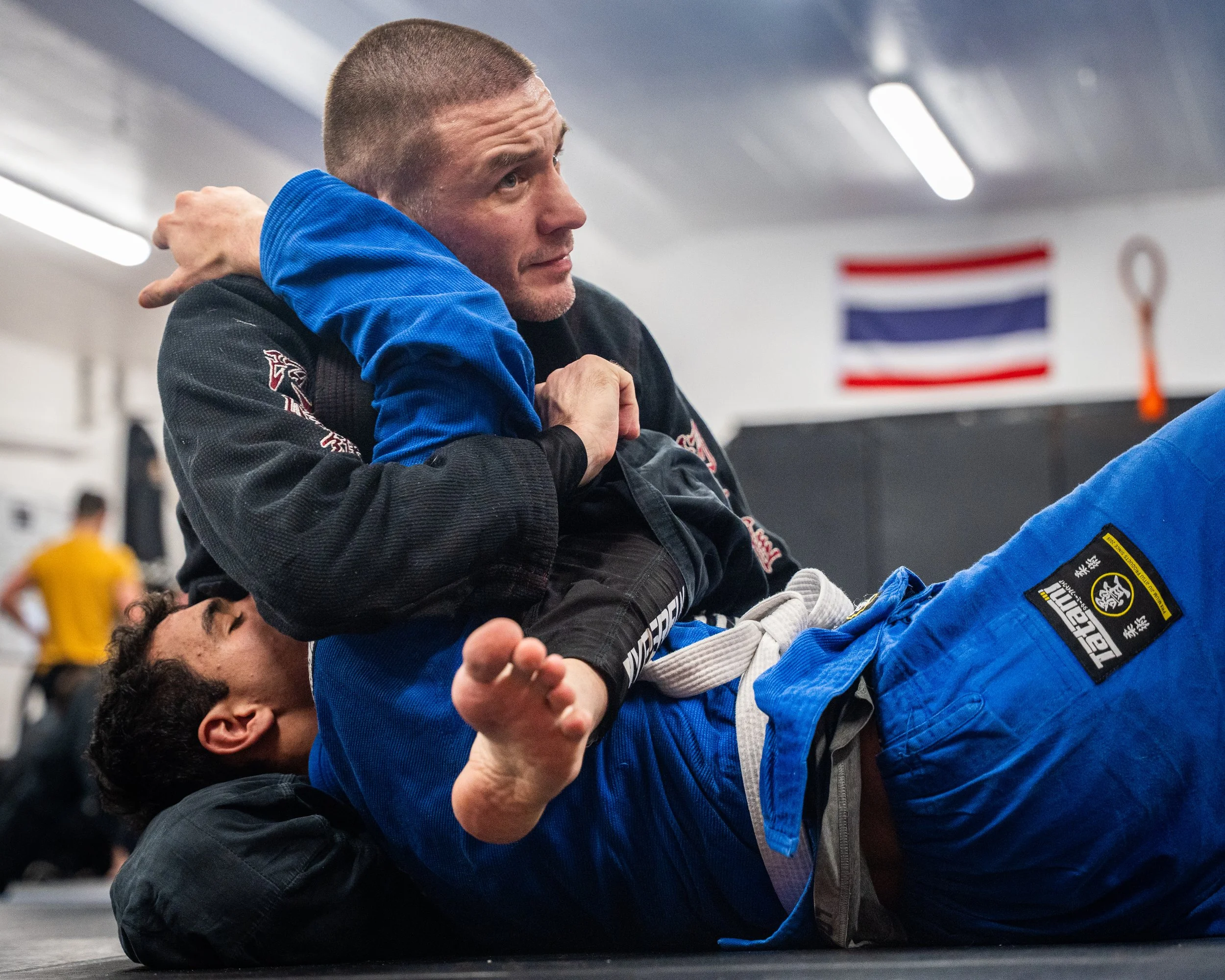 Två personer utövar brasiliansk jiu-jitsu i en träningslokal, den översta personen, en man i en blå gi, är på rygg och den understa personen, en man i svart gi, är ovanpå och håller fast honom.