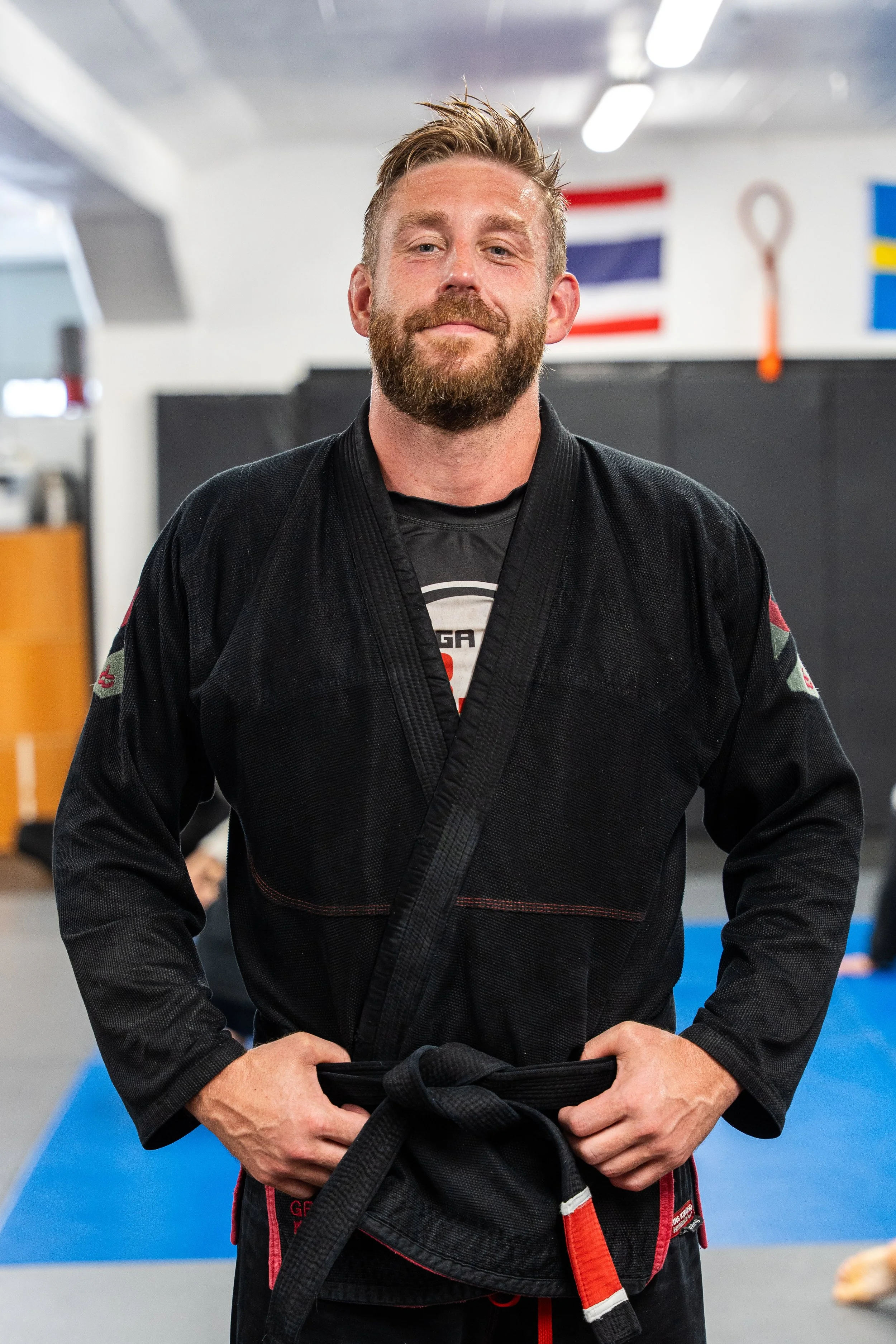 En man i svart jiu-jitsu kimono står i ett träningscenter, håller ett svart bälte i händerna.