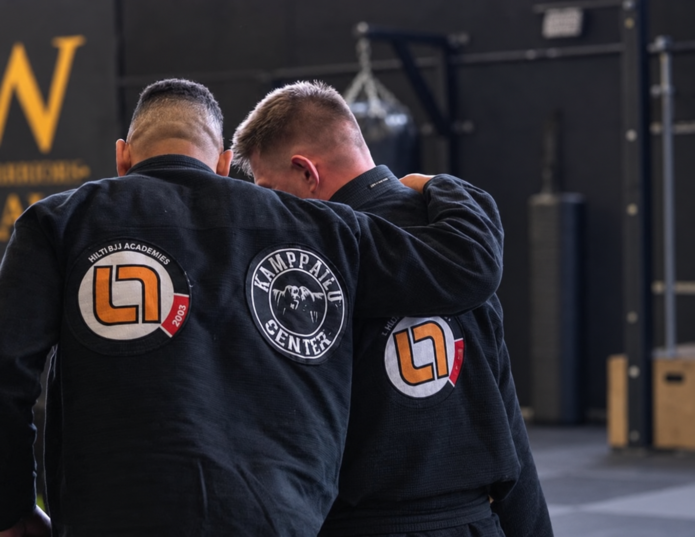 Två personer i jiu-jitsu-gi med patchar som visar kampcenter och ett helsidesmärken, en av dem tröstar den andre.