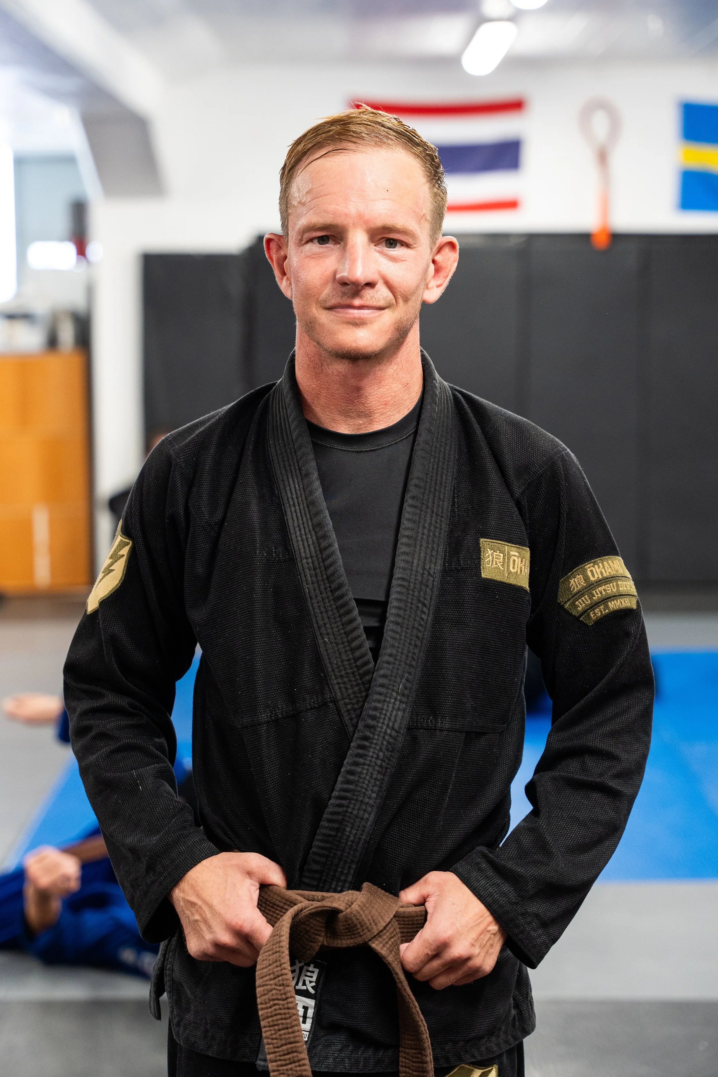 Man i svart jiu-jitsu dräkt med bältet knutet runt midjan, står i ett träningsutrymme. Bakgrund avlägset med en person i blå dräkt och andra träningsredskap, flaggor på väggen.