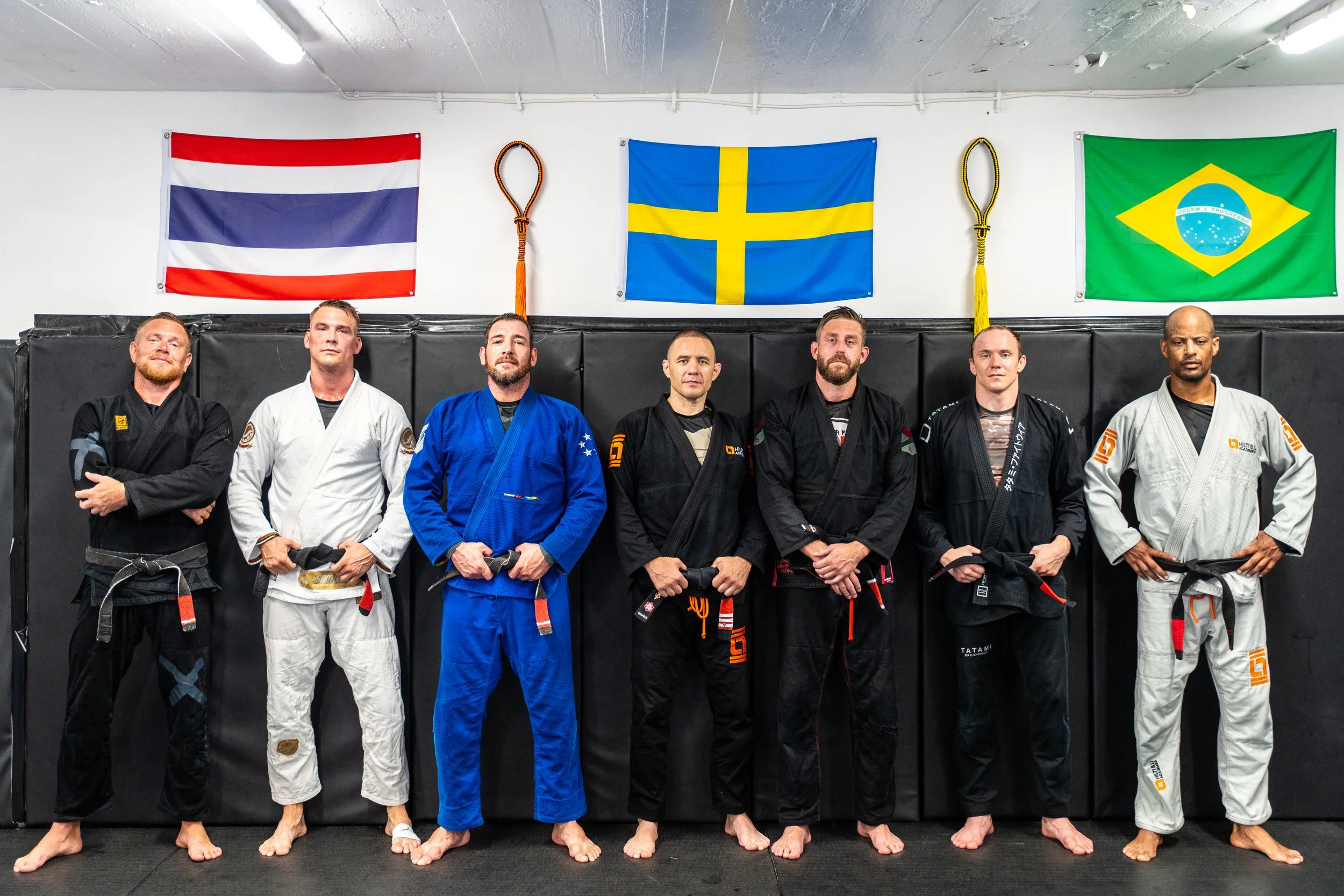 Grupp av sju personer i jujutsu-uniformer som står i rad framför en vit vägg med tre flaggor: Nederländernas, Sveriges och Brasiliens.