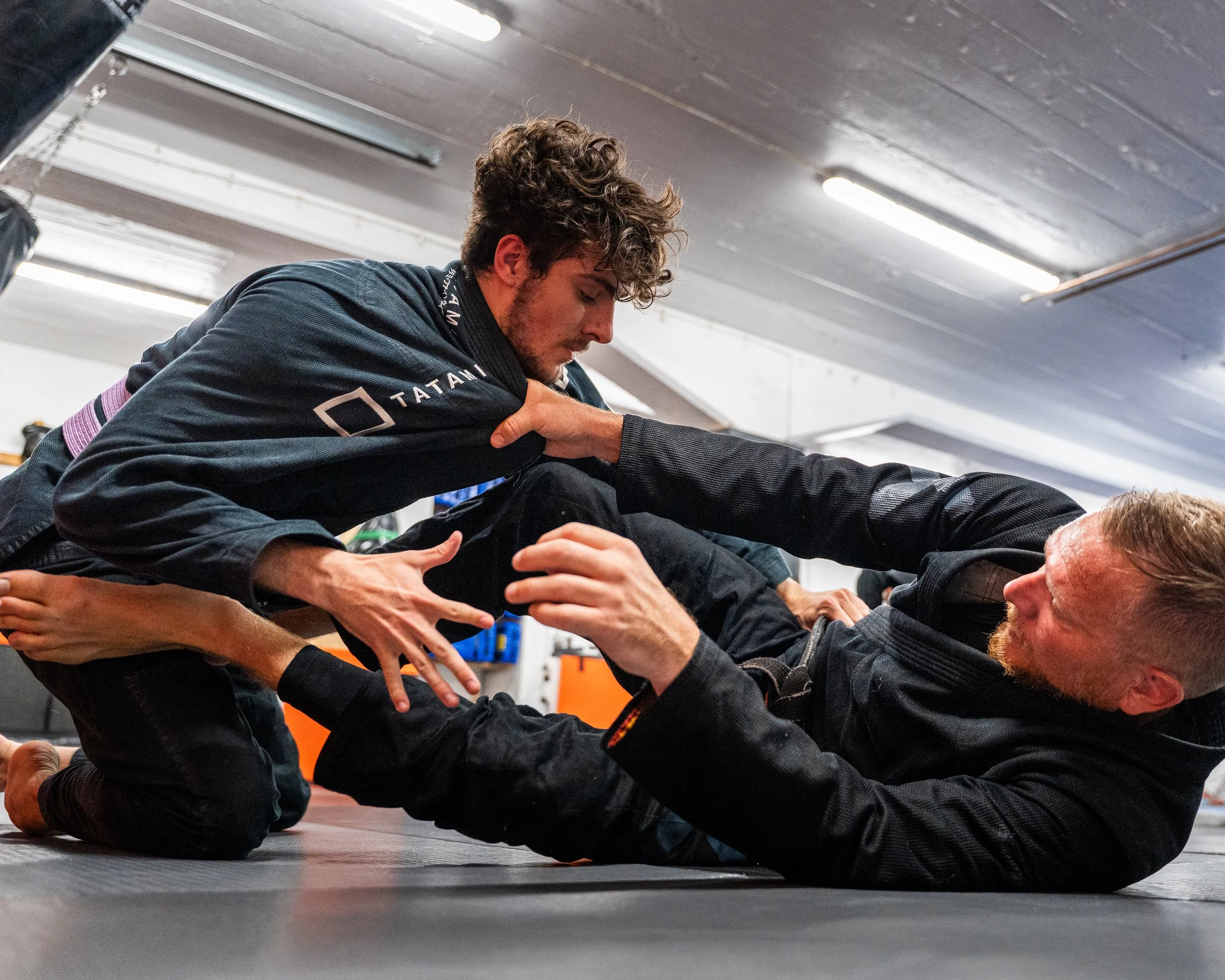 Två personer deltar i jiu-jitsu träning på en matta, den ena personen är en man med lockigt hår, den andra är en man med kort hår, båda är iförda jiu-jitsu dräkter.