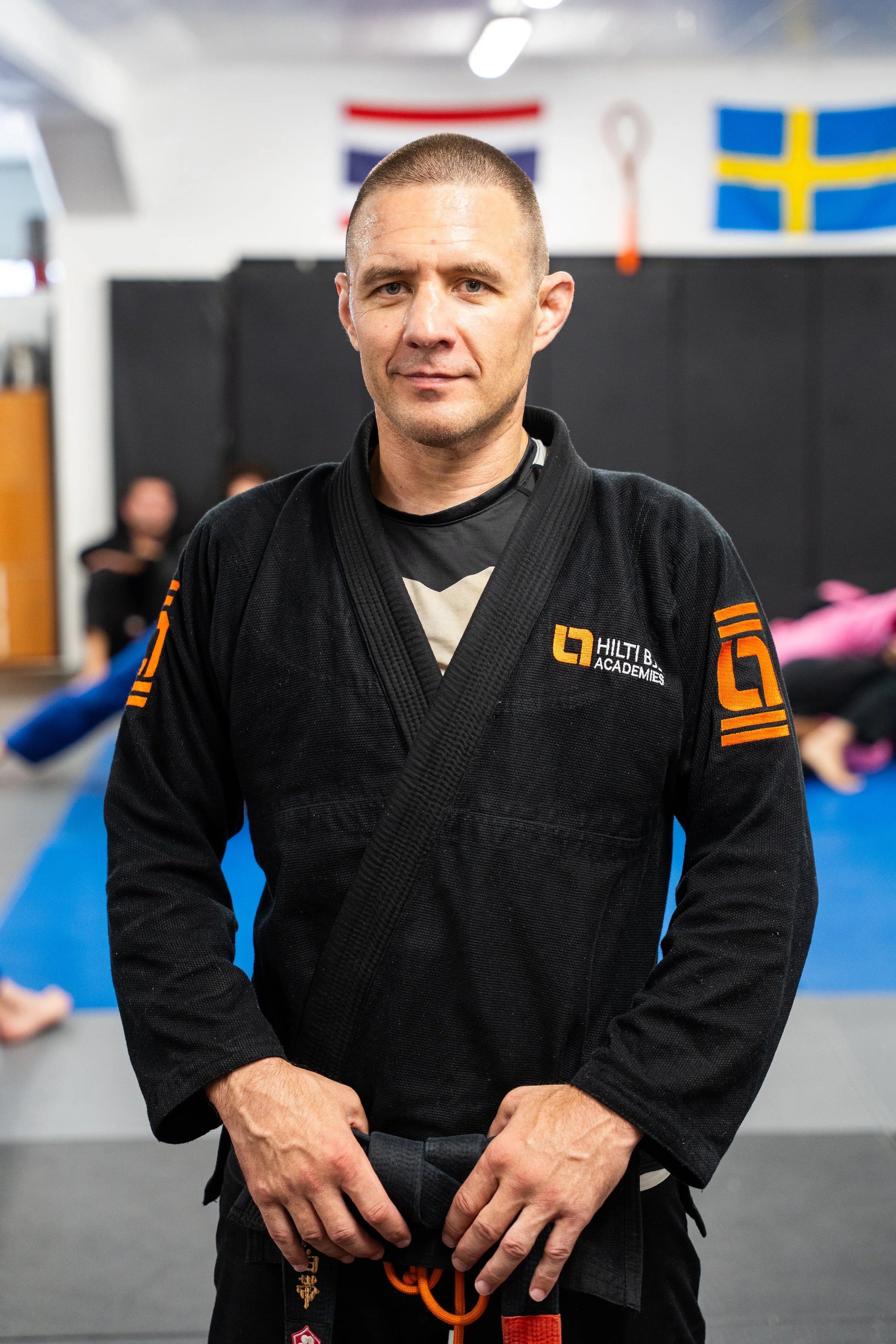 Man i svart jiu-jitsu-dräkt står i en dojon, med flaggor av Norge och Sverige i bakgrunden.
