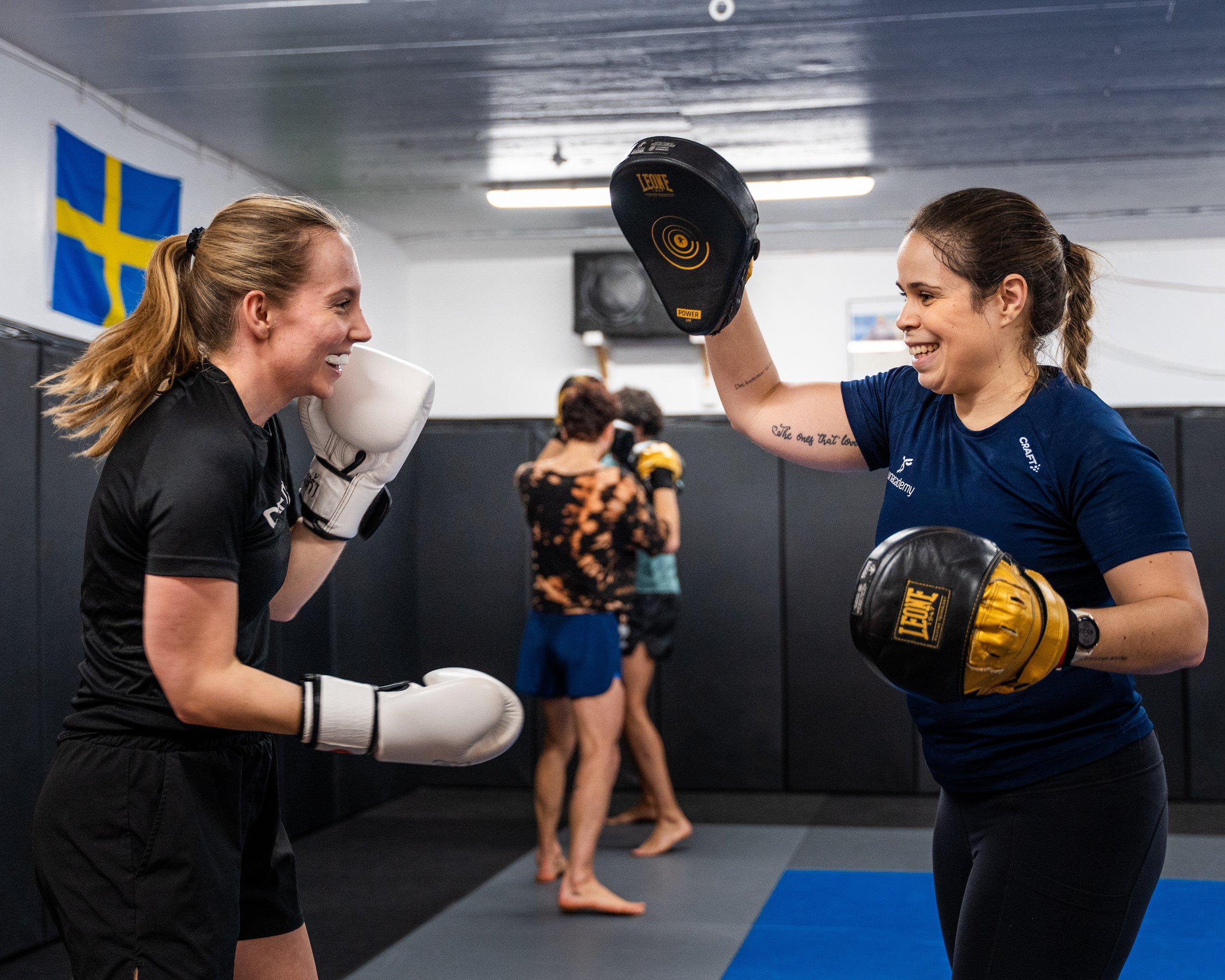 Två kvinnor tränar kampsport i en träningsstudio. Den ena kvinna har boxhandskar och den andra håller en fokusplatta. Båda är glada och ler, en kvinna har håret i en hästsvans och den andra med lockigt hår. En svensk flagga hänger på väggen.