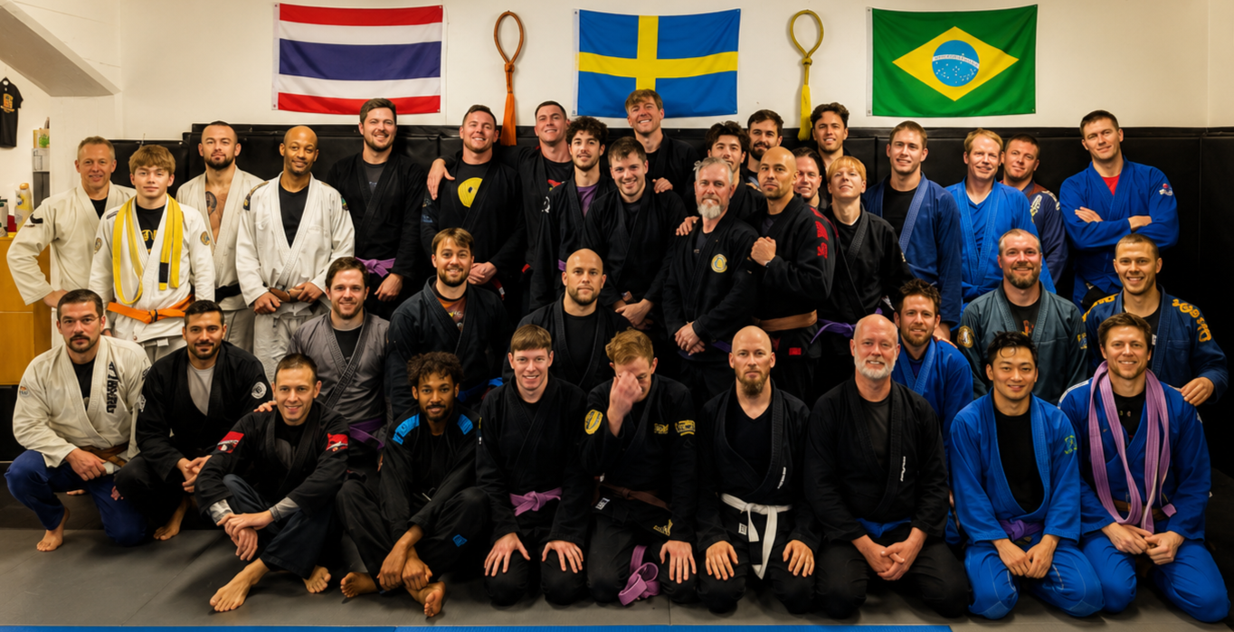 En grupp personer i jiu-jitsu-uniformer står tillsammans i en träningslokal, med flaggor från Thailand, Sverige och Brasilien på väggen bakom.