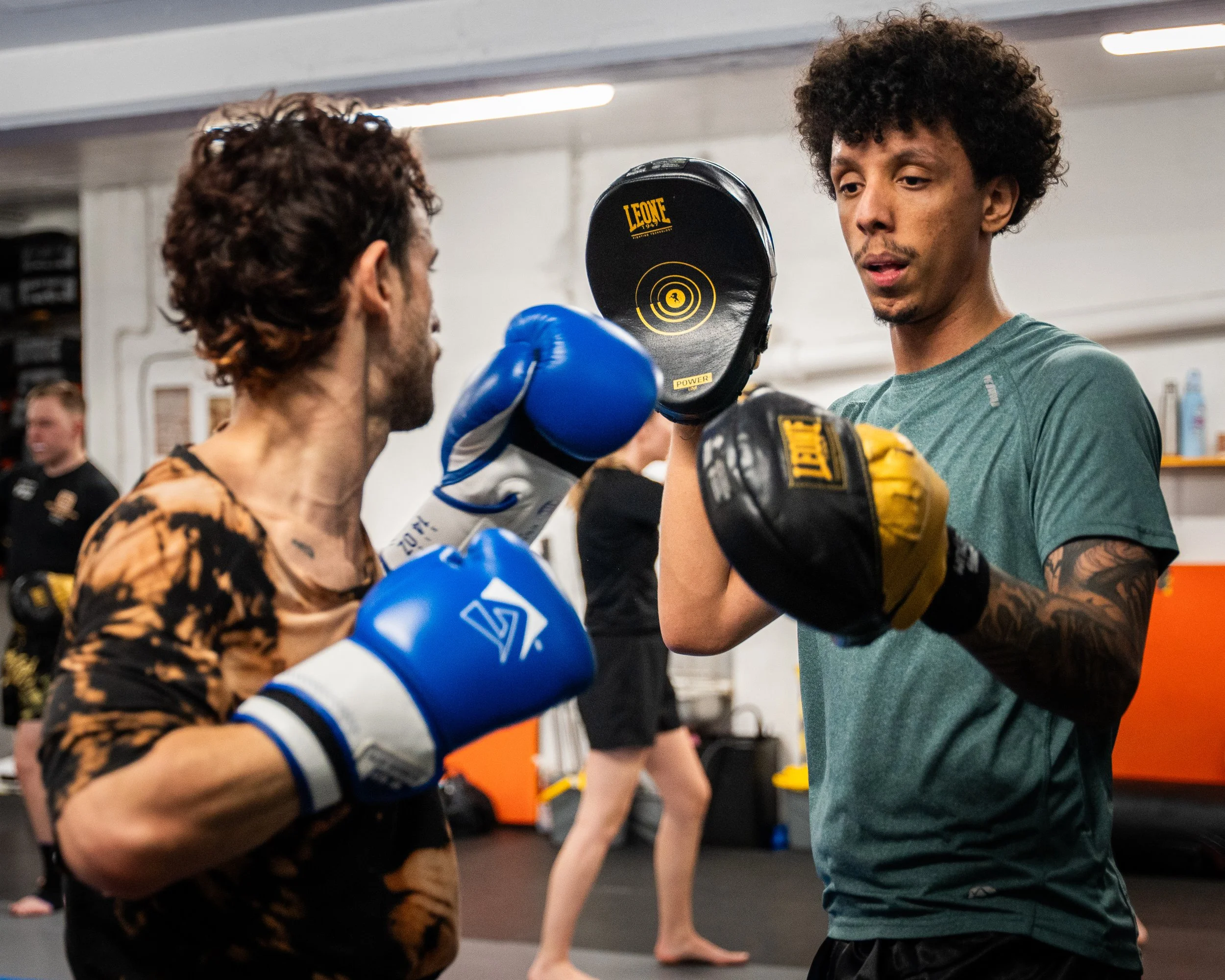 Två personer tränar boxning i en boxinghall, en av dem bär blå handskar och den andra bär gula handskar. De är fokuserade och tränar tillsammans.