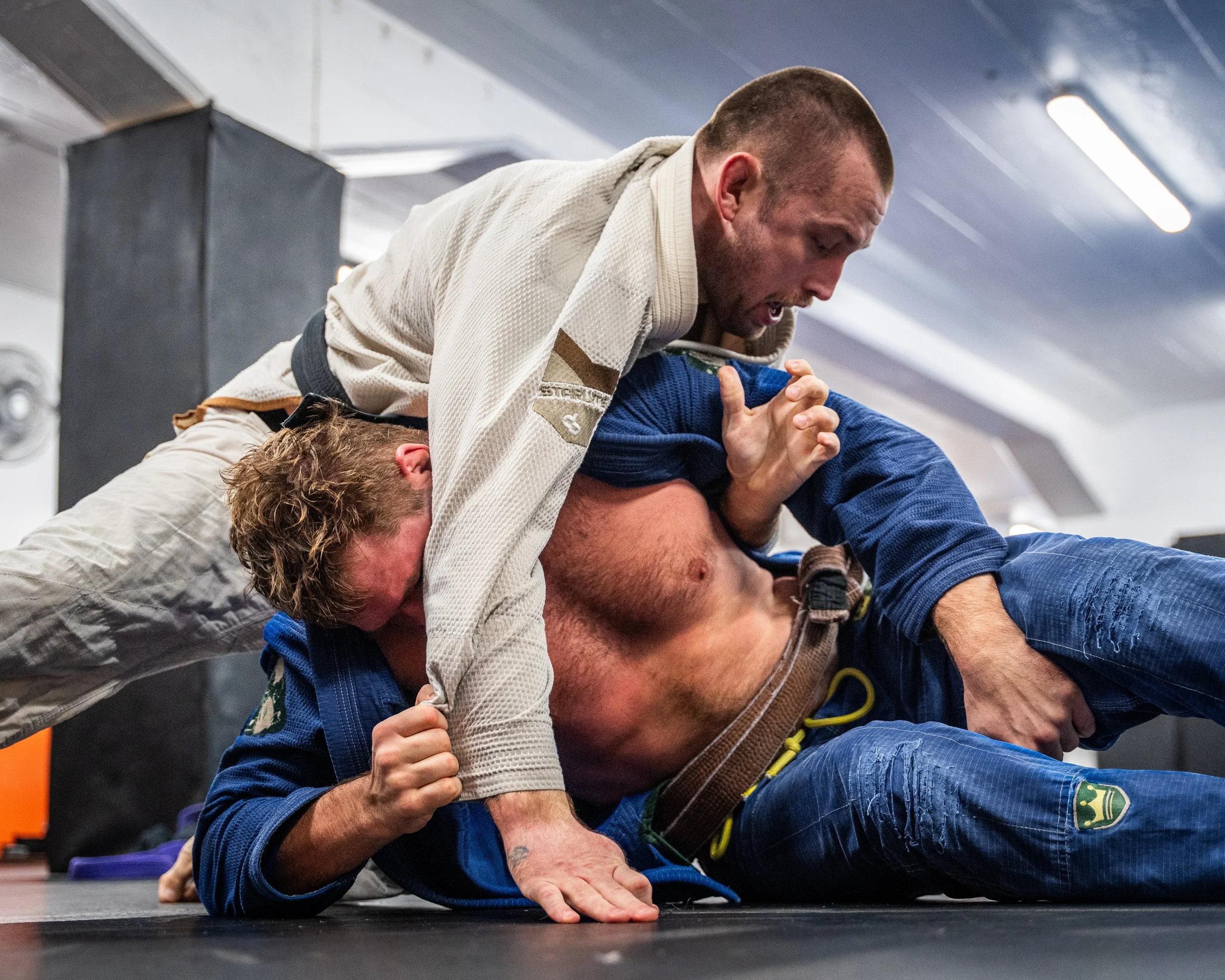 Två personer tränar jiu jitsu på golvet, en person i vit gi håller fast en person i blått gi under en träningsövning.