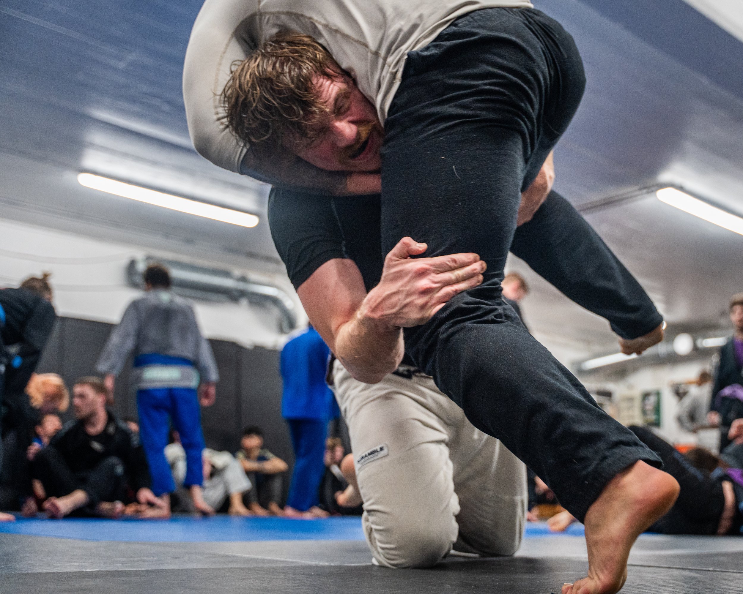 Två personer tävlar i brasiliansk jiu-jitsu, en i en svart dräkt kämpar på golvet medan den andra, i en beige dräkt, håller fast den och fastställer en position i en träningslokal.