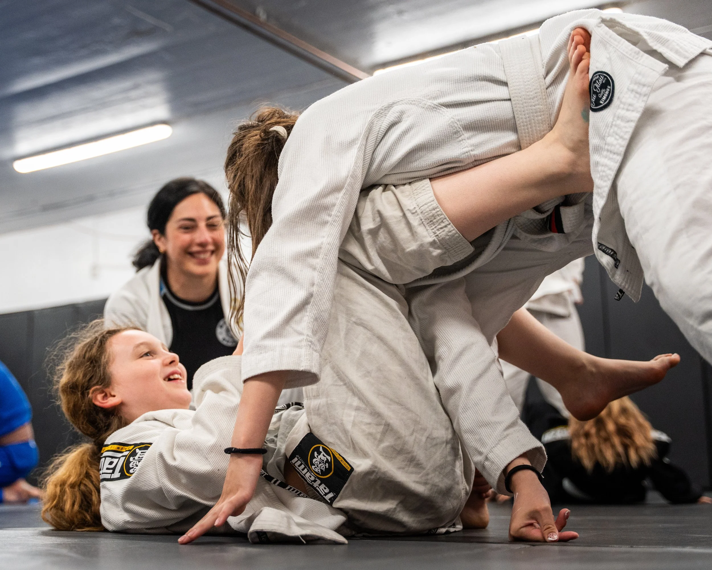 Två personer i judodräkter som utför ett gruppövning eller sport i ett träningshall, med andra personer i bakgrunden.
