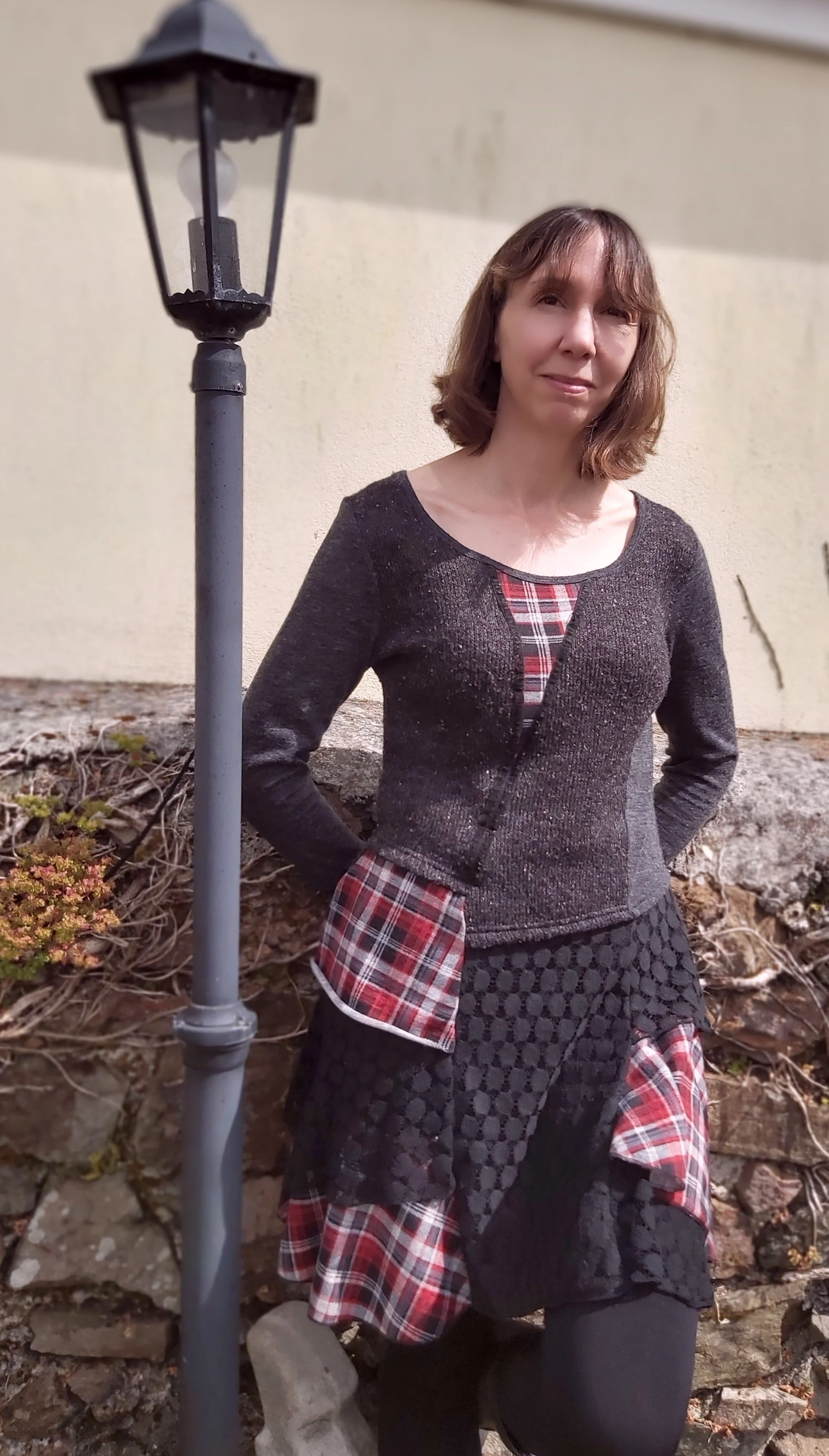 A woman with shoulder-length brown hair standing outdoors near a lamp post, wearing a black cardigan with red plaid accents and black patterned pants, with a beige wall behind her.