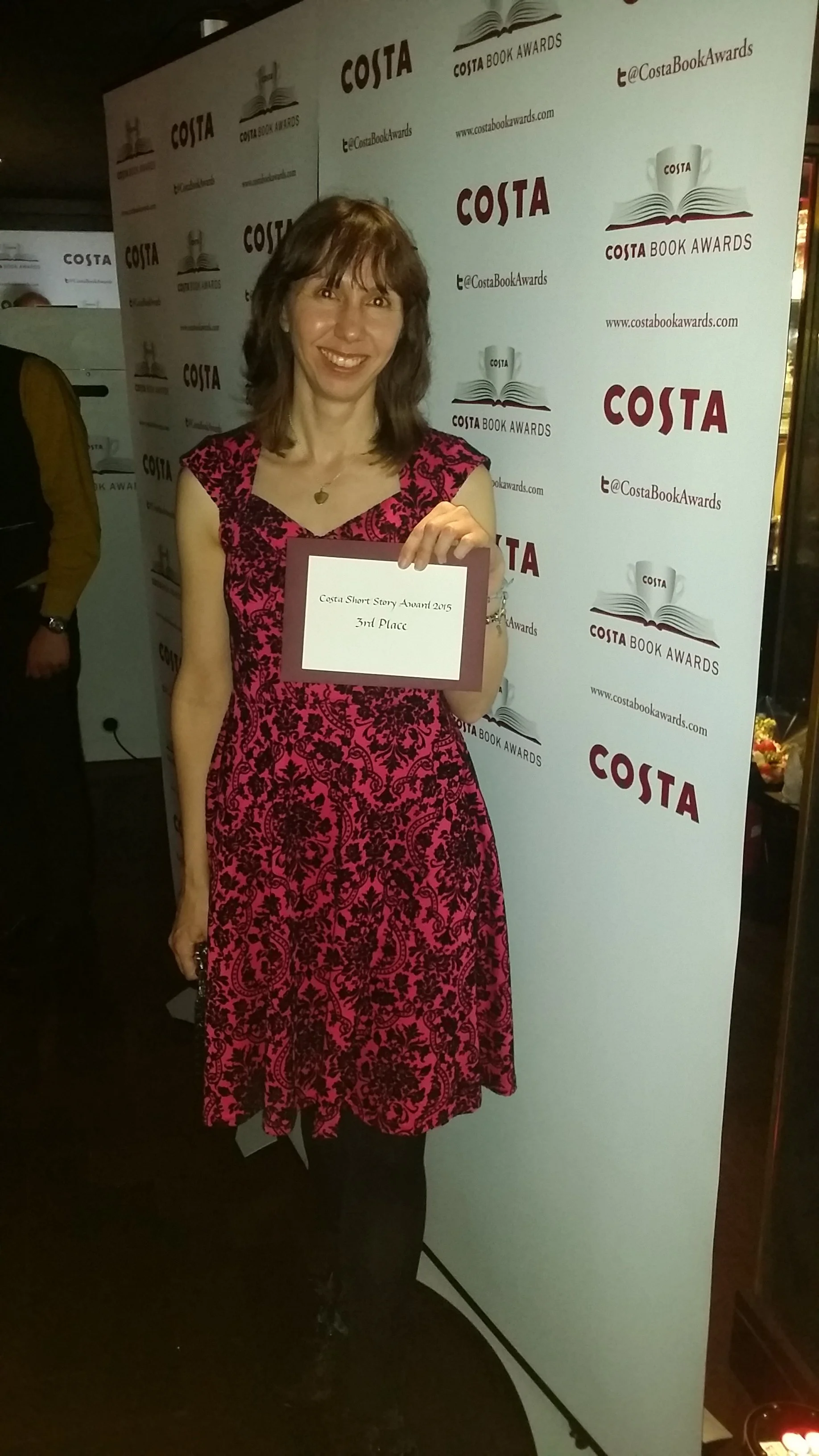 A woman in a pink and black patterned dress smiling and holding an award certificate at the Costa Book Awards event.