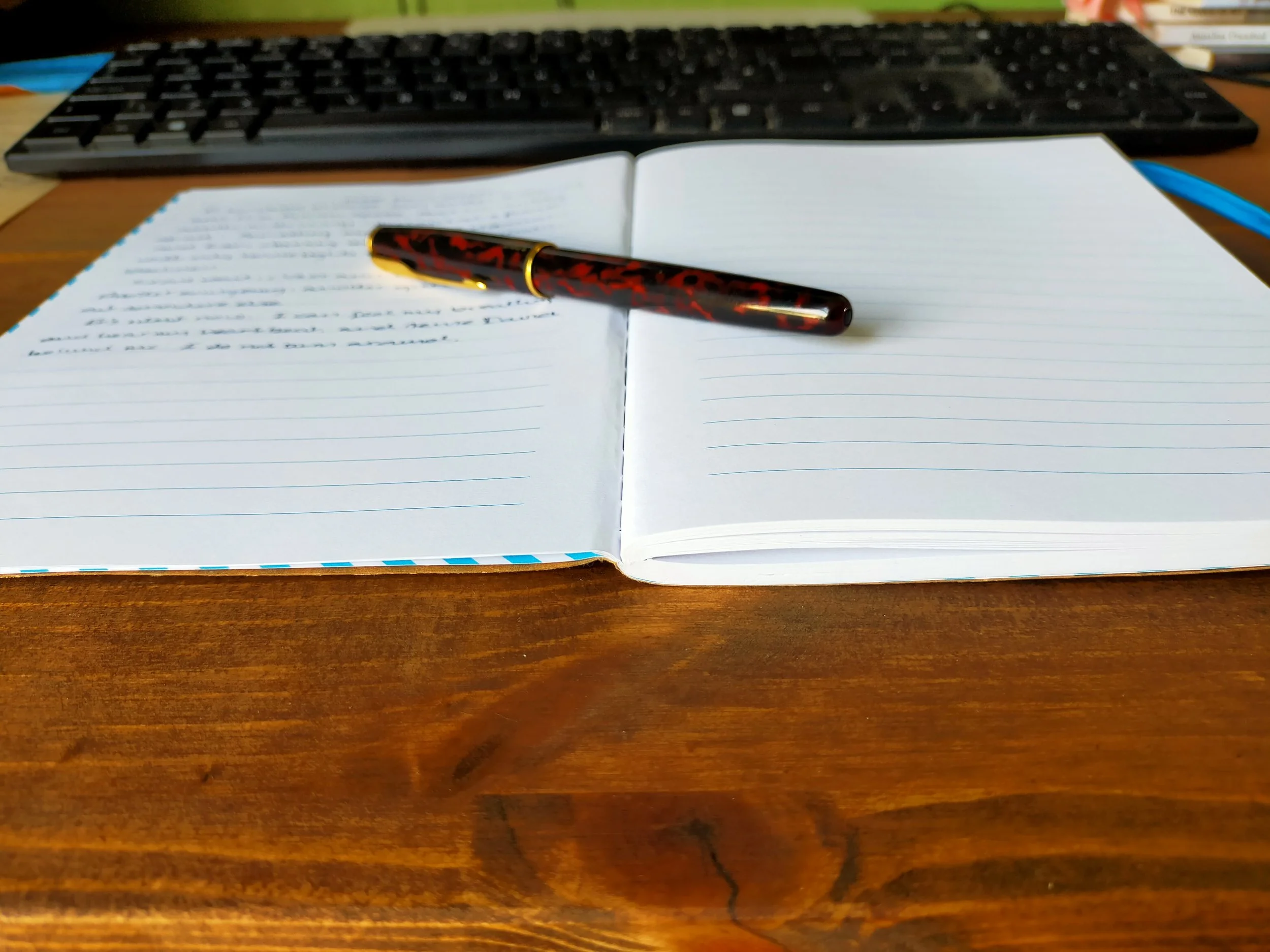 An open notebook on a wooden desk with blue lines and some handwritten notes, a black and red patterned pen resting on the page, and a computer keyboard in the background.