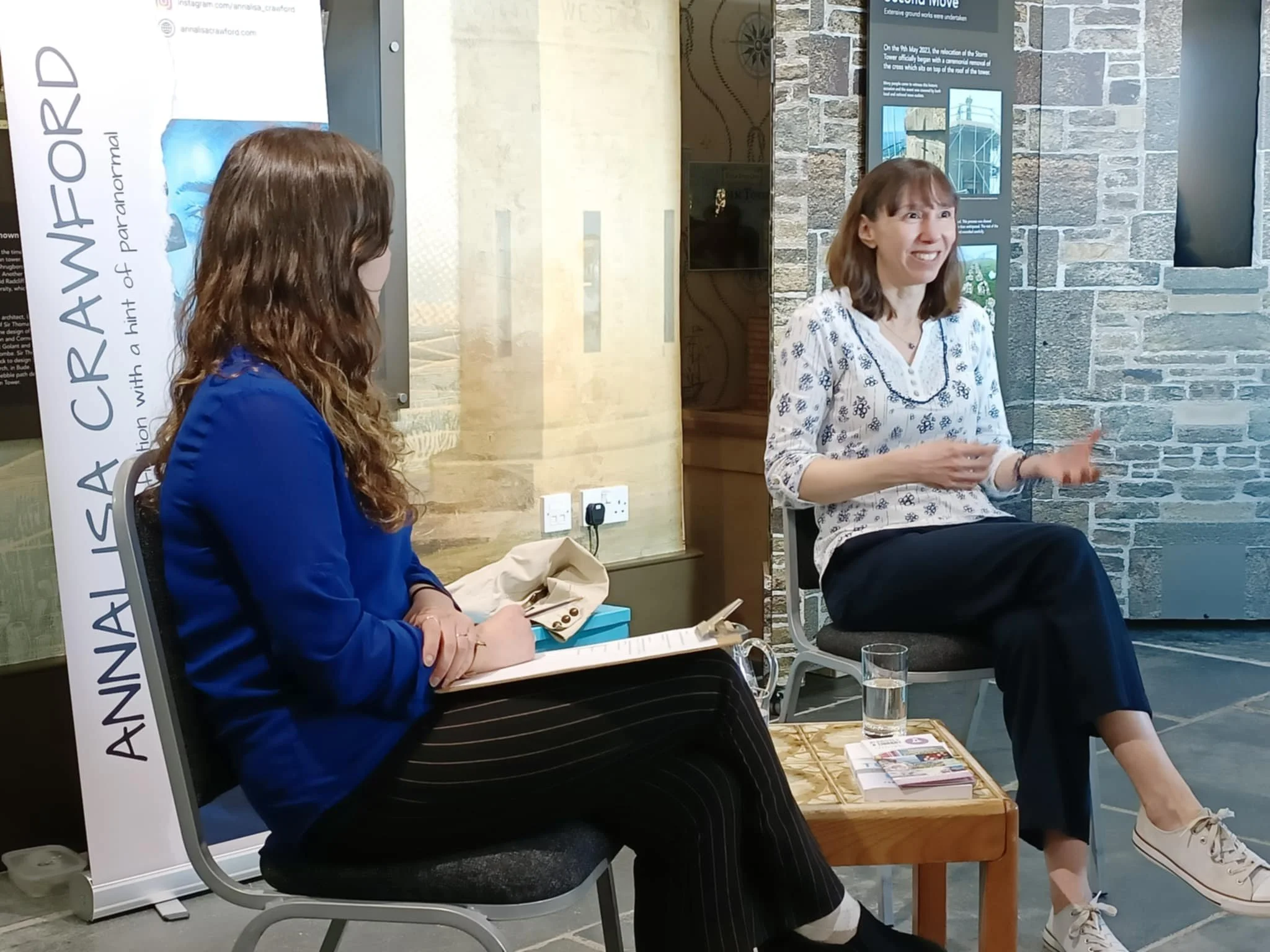 Two women seated and engaged in conversation in a room with stone and wood walls. One woman has light brown hair and is smiling, wearing a white blouse with black patterns. The other woman has long, wavy brown hair, and is wearing a blue blazer. There are informational posters and brochures on a table in front of them.