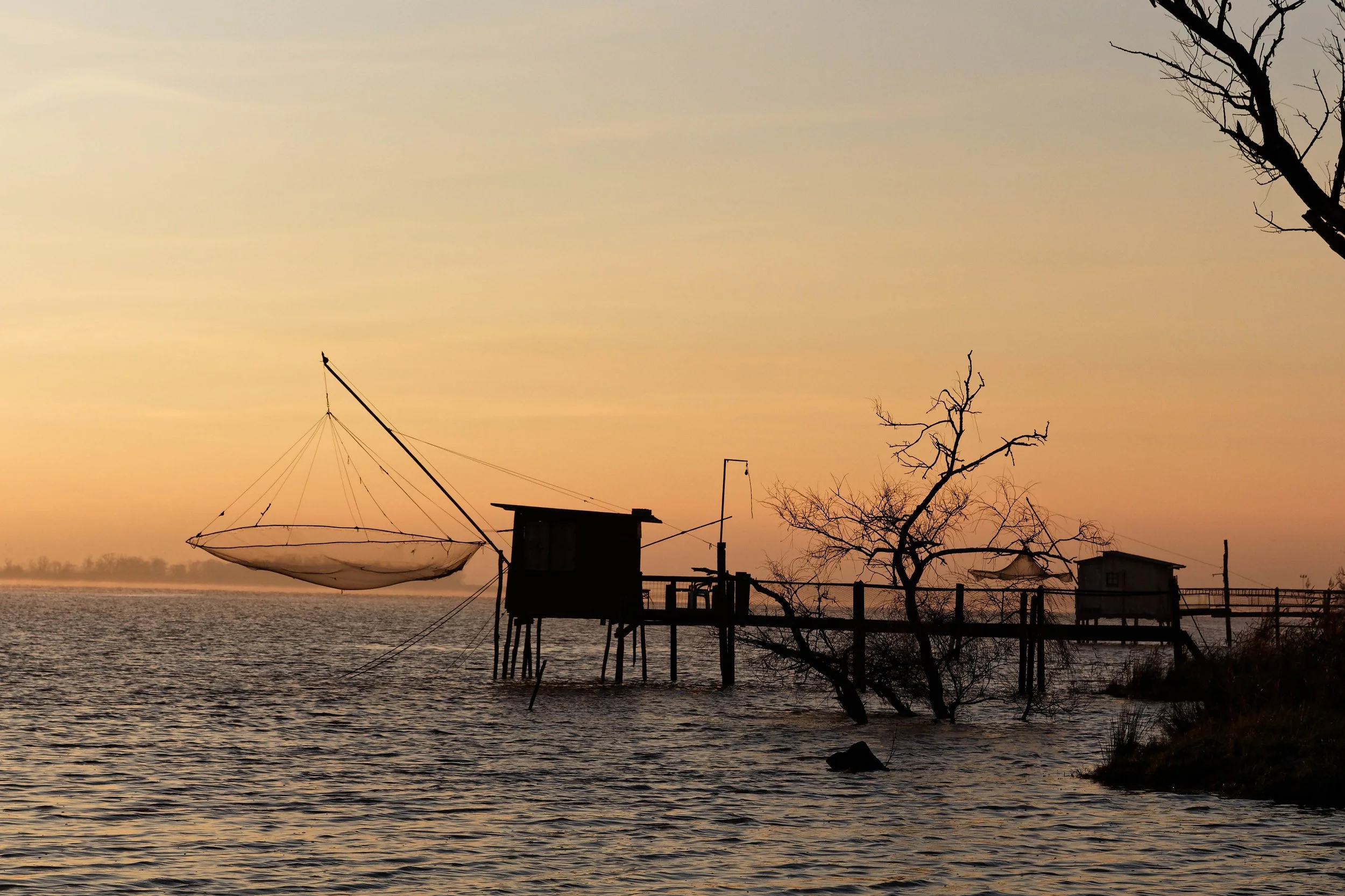Scène de coucher de soleil avec un horizon calme, des arbres nus, une maison en bois sur pilotis au bord de l'eau, un filet de pêche suspendu à une structure, et un ciel aux teintes orangées.