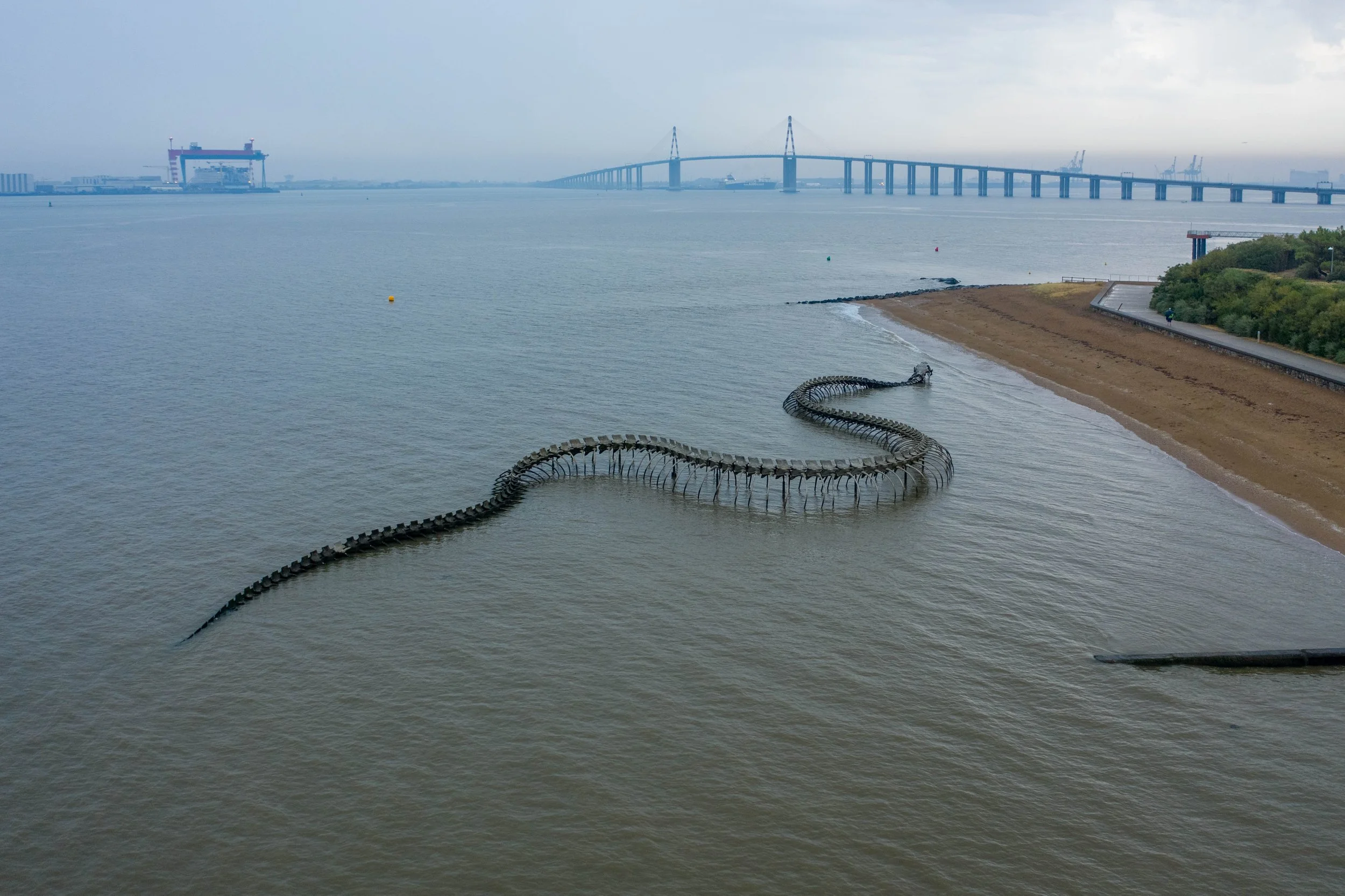 Une vue d'un pont suspendu au-dessus de l'eau, avec une plage et un chemin piéton à droite. Il y a une structure en spirale dans l'eau près de la plage. En arrière-plan, des grues industrielles et des bâtiments.
