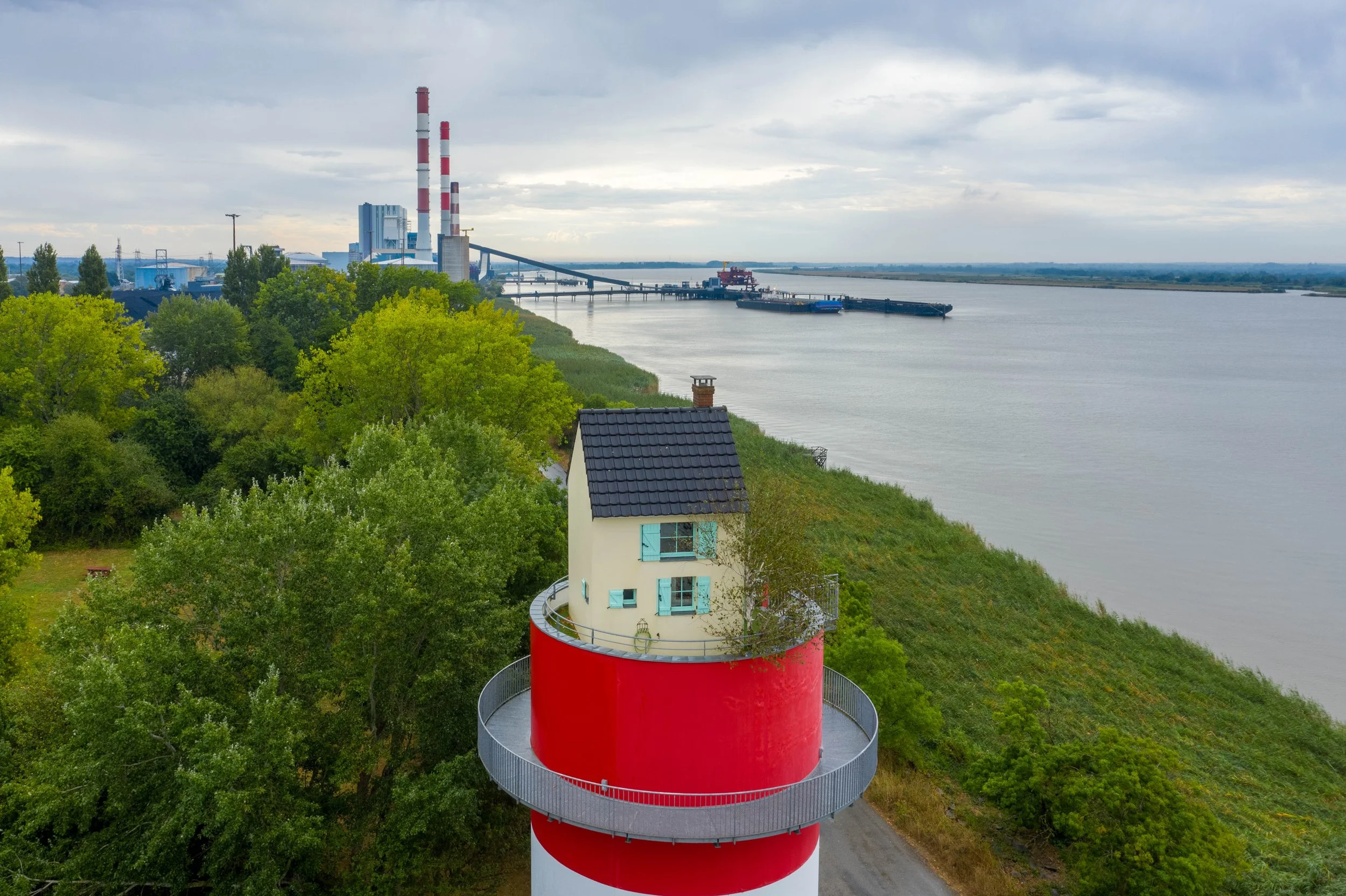 Un phare rouge et blanc en spirale avec une maison à fenêtres bleues, entouré d'arbres verts, situé près d'une rivière ou d'un lac, avec une usine au loin avec des cheminées.