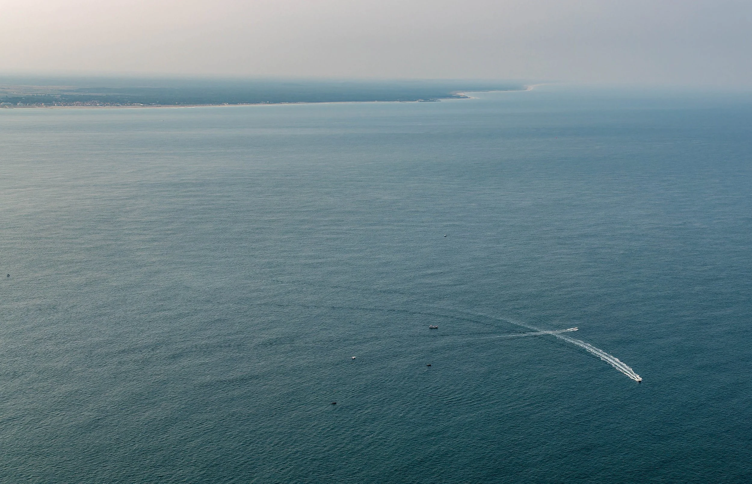 Vue aérienne de la mer avec plusieurs bateaux laissant des traces dans l'eau, et une côte au loin sous un ciel brumeux.