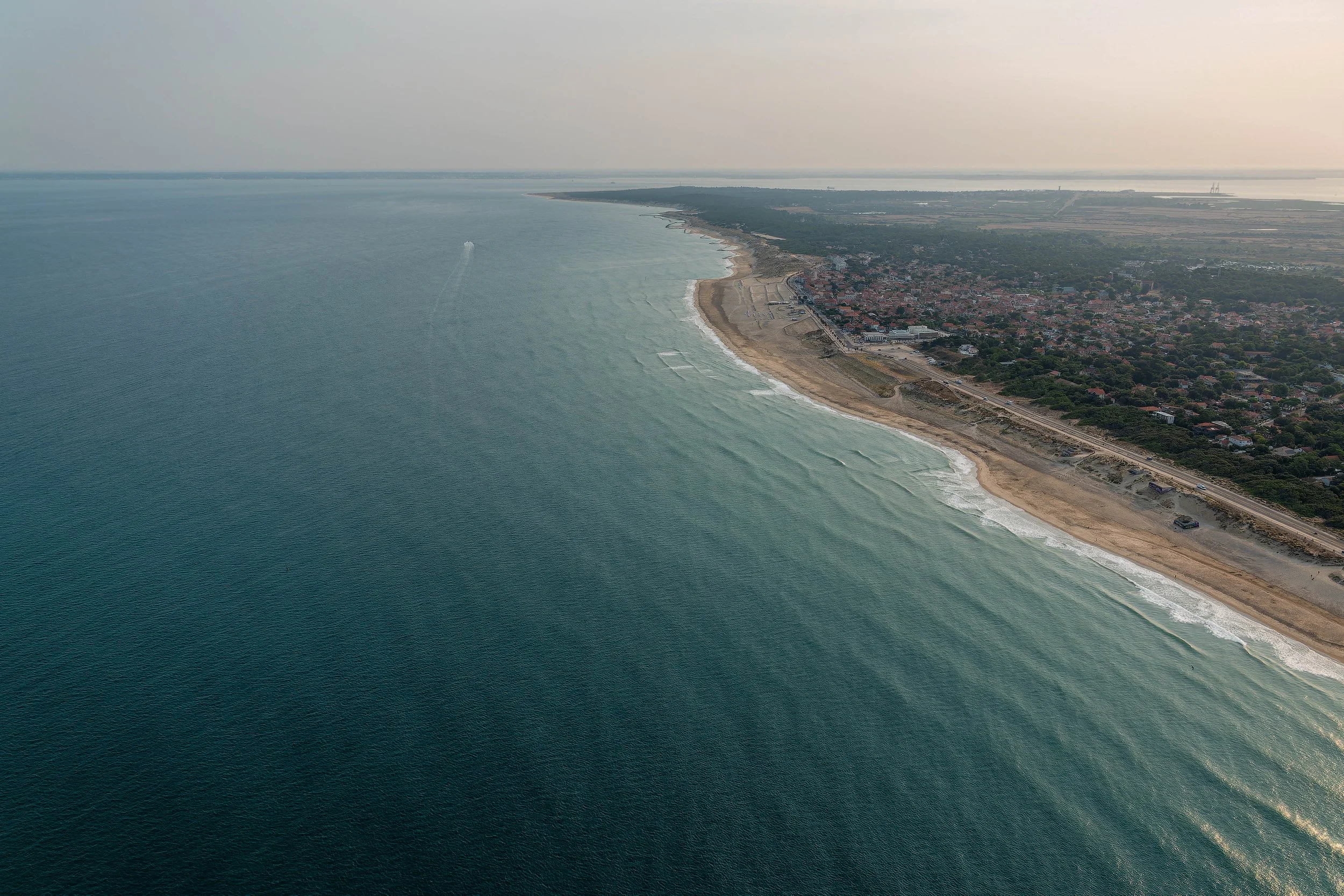 Vue aérienne d'une côte avec une plage de sable et un quartier résidentiel, en arrière-plan des terres agricoles et des zones industrielles.