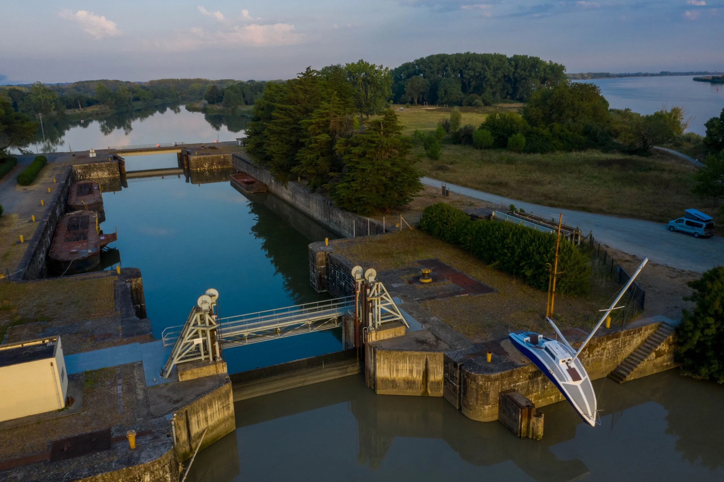 Vue aérienne d'un canal de dérivation d'eau avec des structures métalliques et un bateau amarré, entouré d'arbres et d'une nature paisible, avec un fleuve en arrière-plan.