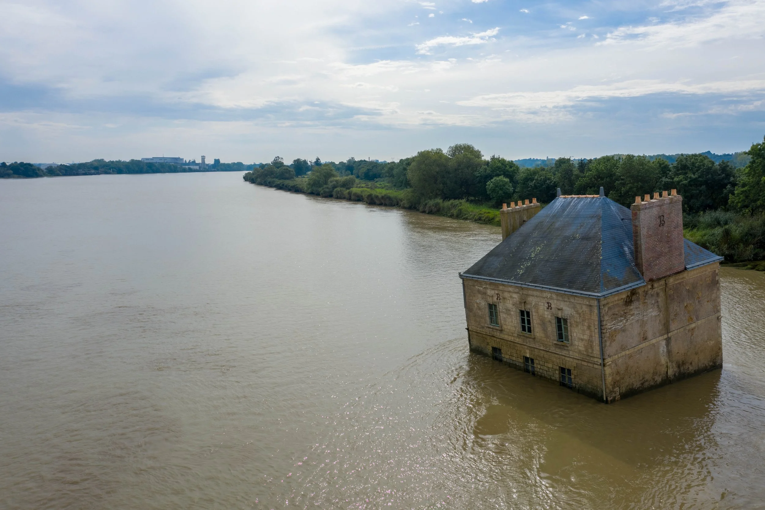 Une vieille maison en pierre située en bord de rivière, avec un toit en tuiles et des cheminées, entourée de verdure et d'arbres, sous un ciel partiellement nuageux.