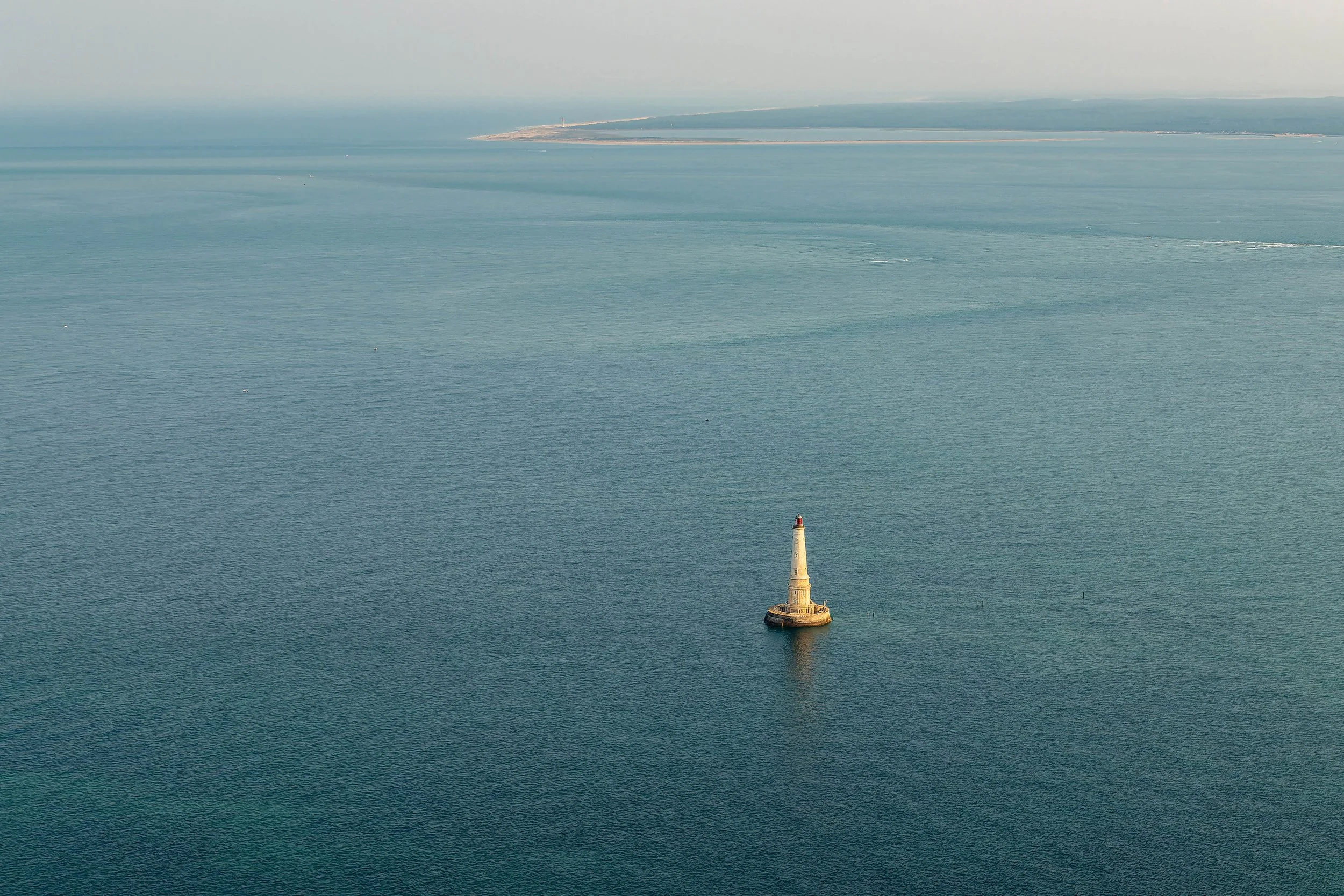 Un phare blanc avec une tête rouge situé dans la mer, avec une île éloignée au fond et le ciel clair.