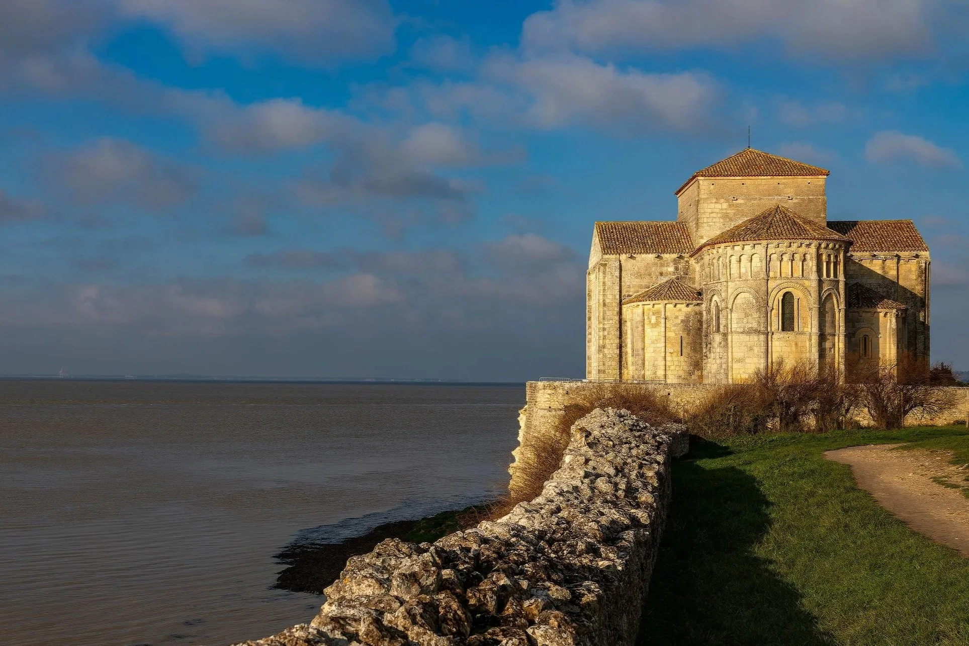 Une ancienne église en pierre située au bord de la mer sous un ciel nuageux avec du soleil éclairant le bâtiment.