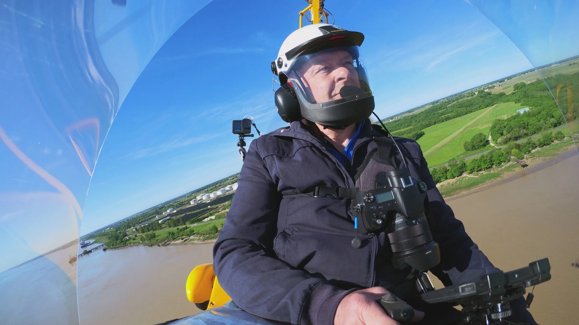 Pilote d'hélicoptère en vol au-dessus d'une rivière traversée par un pont, avec des paysages verts et champêtres en arrière-plan, portant un casque avec visière et un appareil photo suspendu autour du cou.