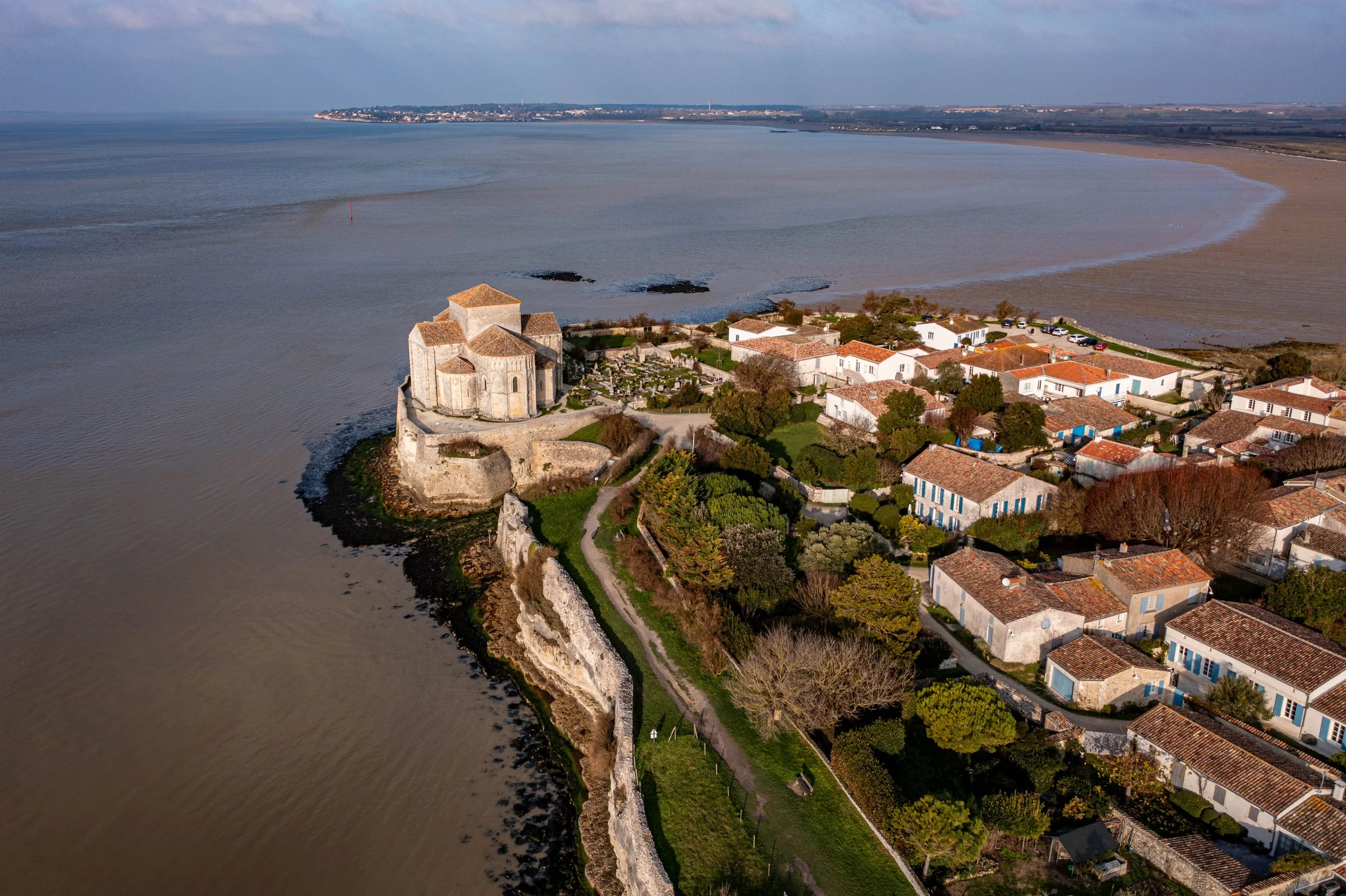 Une vue aérienne d'un village côtier avec une vieille église en pierre et des maisons aux toits en tuiles rouges, entouré de nature verdoyante et de plages, avec la mer au premier plan et une côte lointaine en arrière-plan.