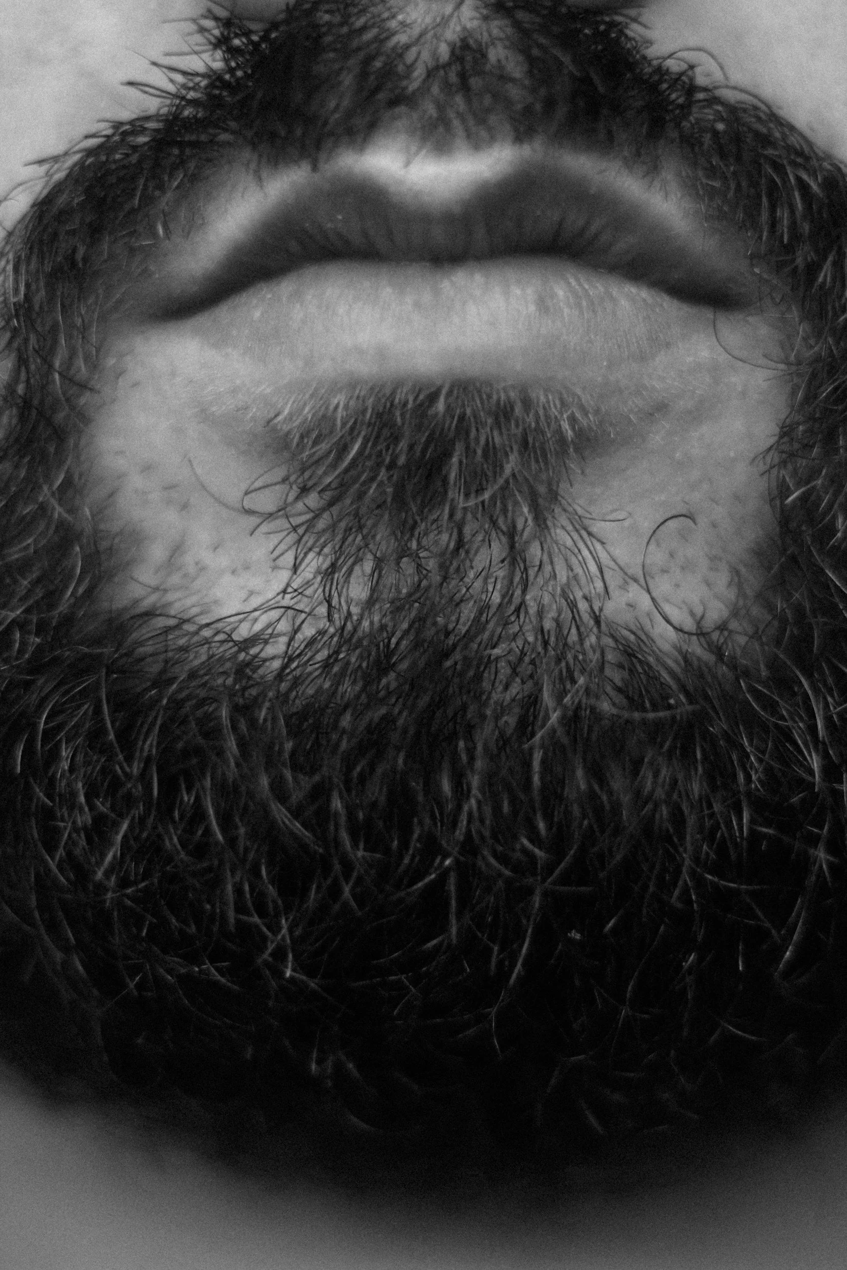 Close-up black and white photo of a person's beard and mustache, with slightly curly hair above.