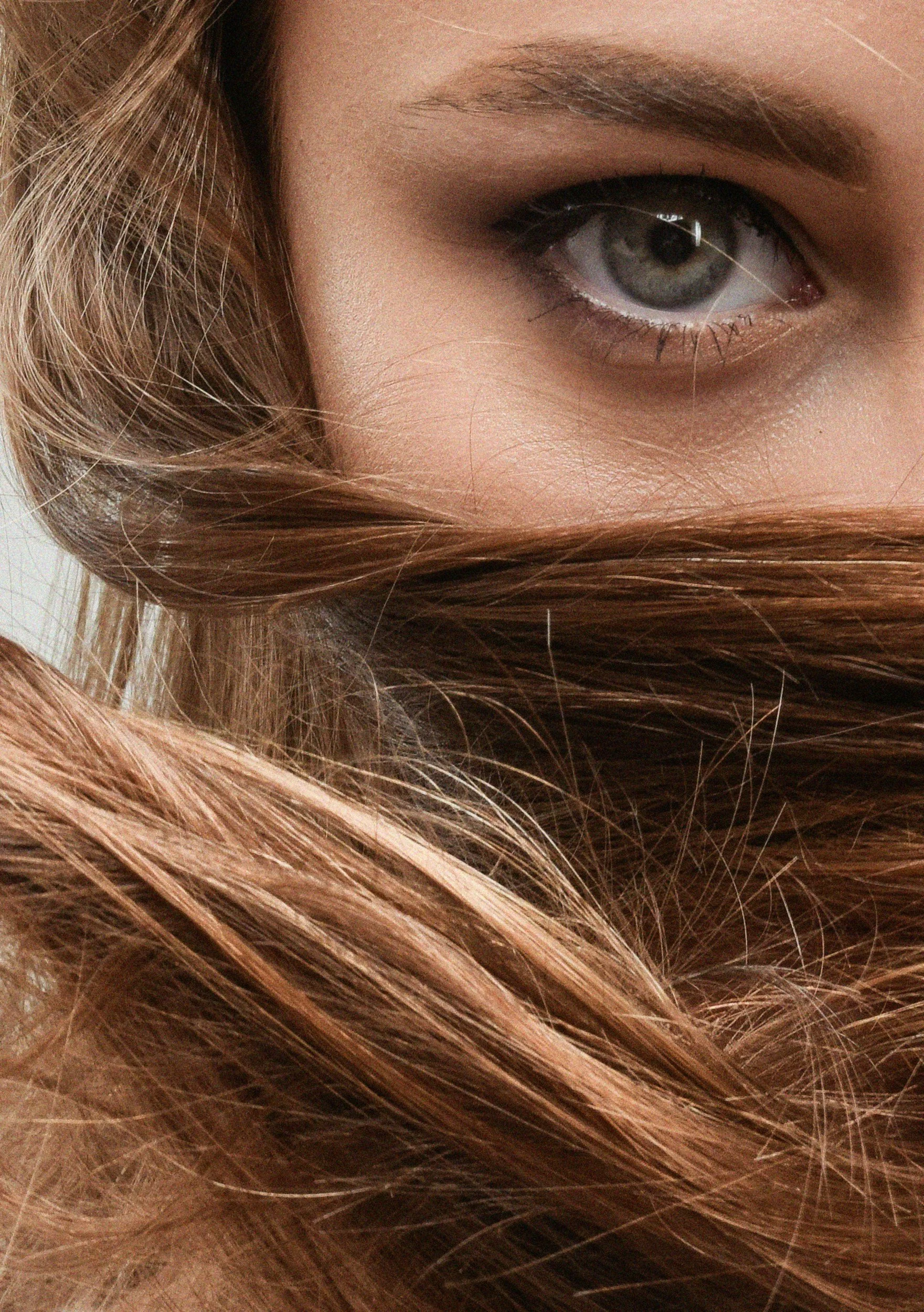Close-up of a person's eye with lengthy, layered light brown hair partially covering the face, showing a well-defined eyebrow, gray eye with detailed eyelashes, and surrounding skin.