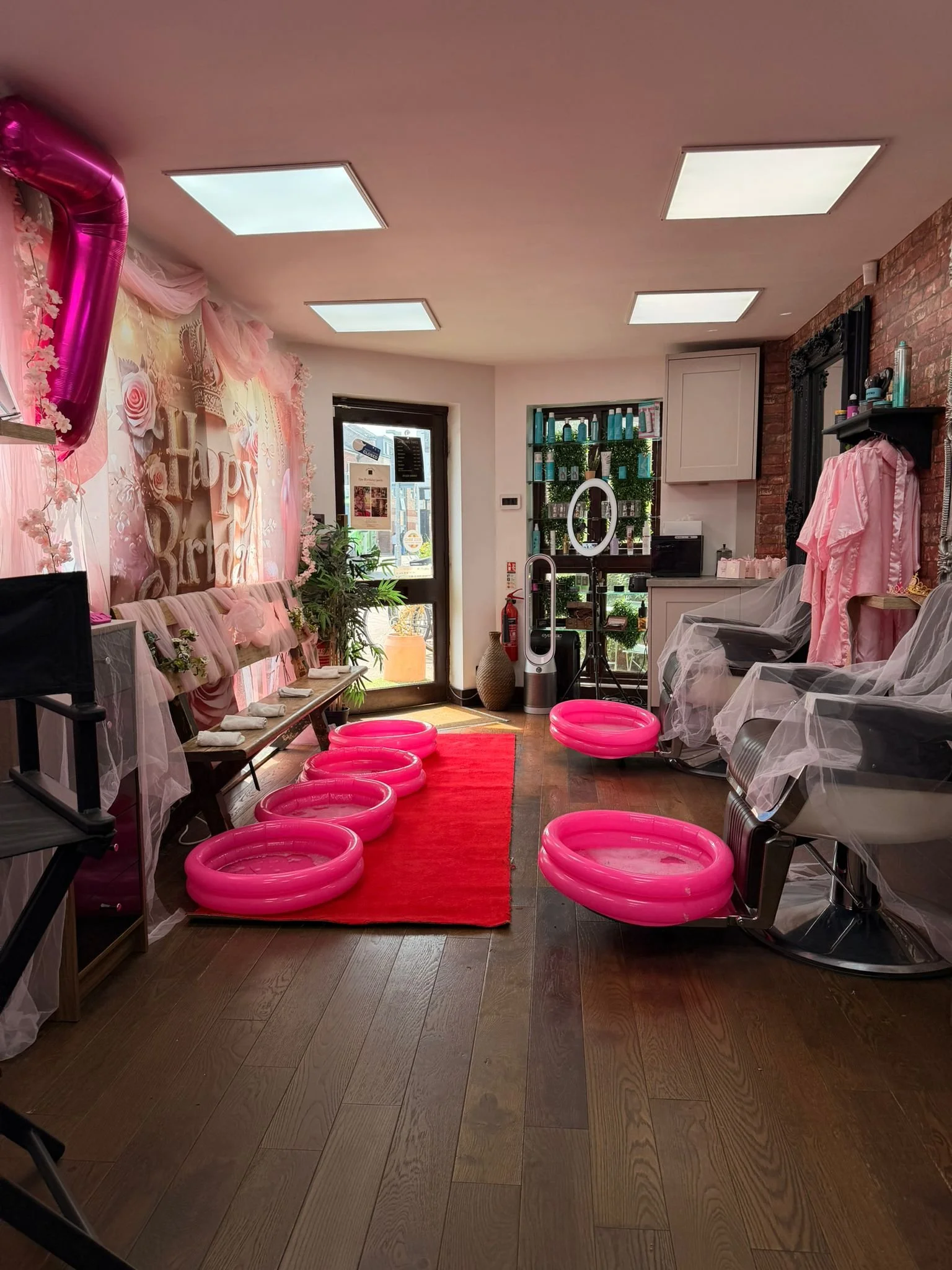 Interior of a decorated event space with pink balloons and drapery, pink rings on the floor, a red carpet, and chairs covered with plastic for a celebration.
