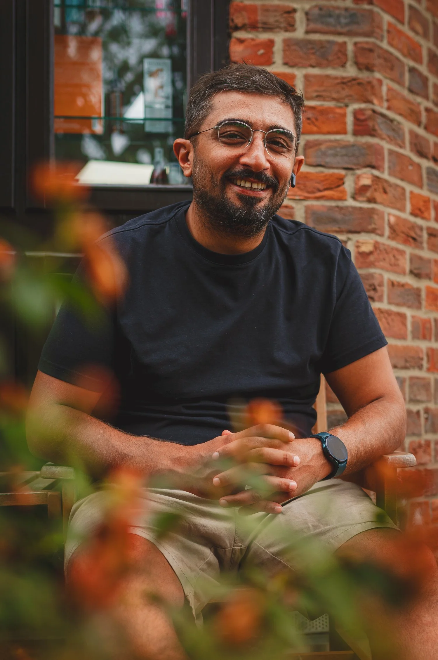 A man with short brown hair, glasses, and a beard smiling and sitting outdoors near a brick wall. He is wearing a black t-shirt, khaki shorts, and a smartwatch.