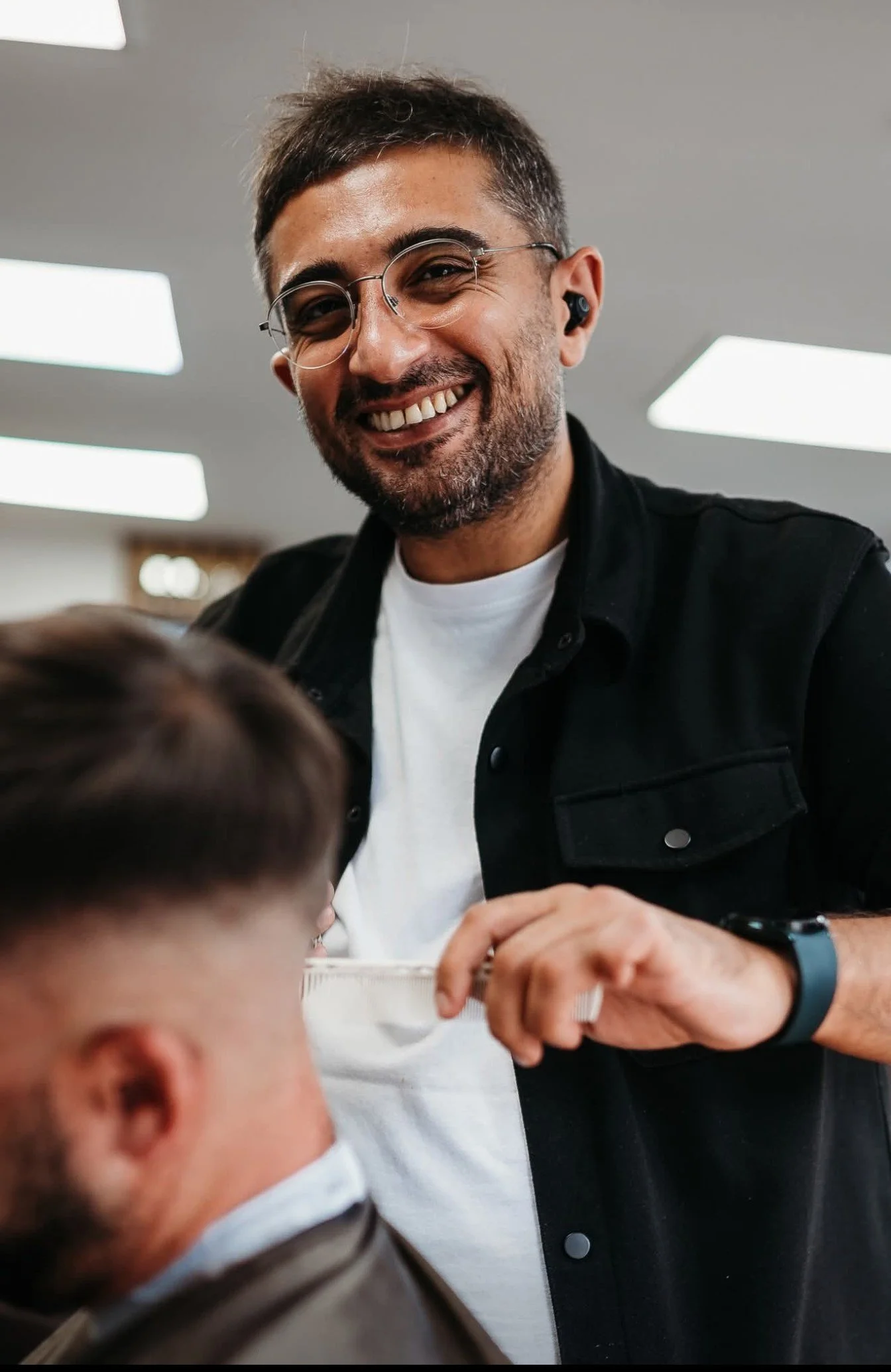 A man with dark hair, glasses, and a beard, smiling while interacting with a child in a classroom setting, with fluorescent ceiling lights visible.