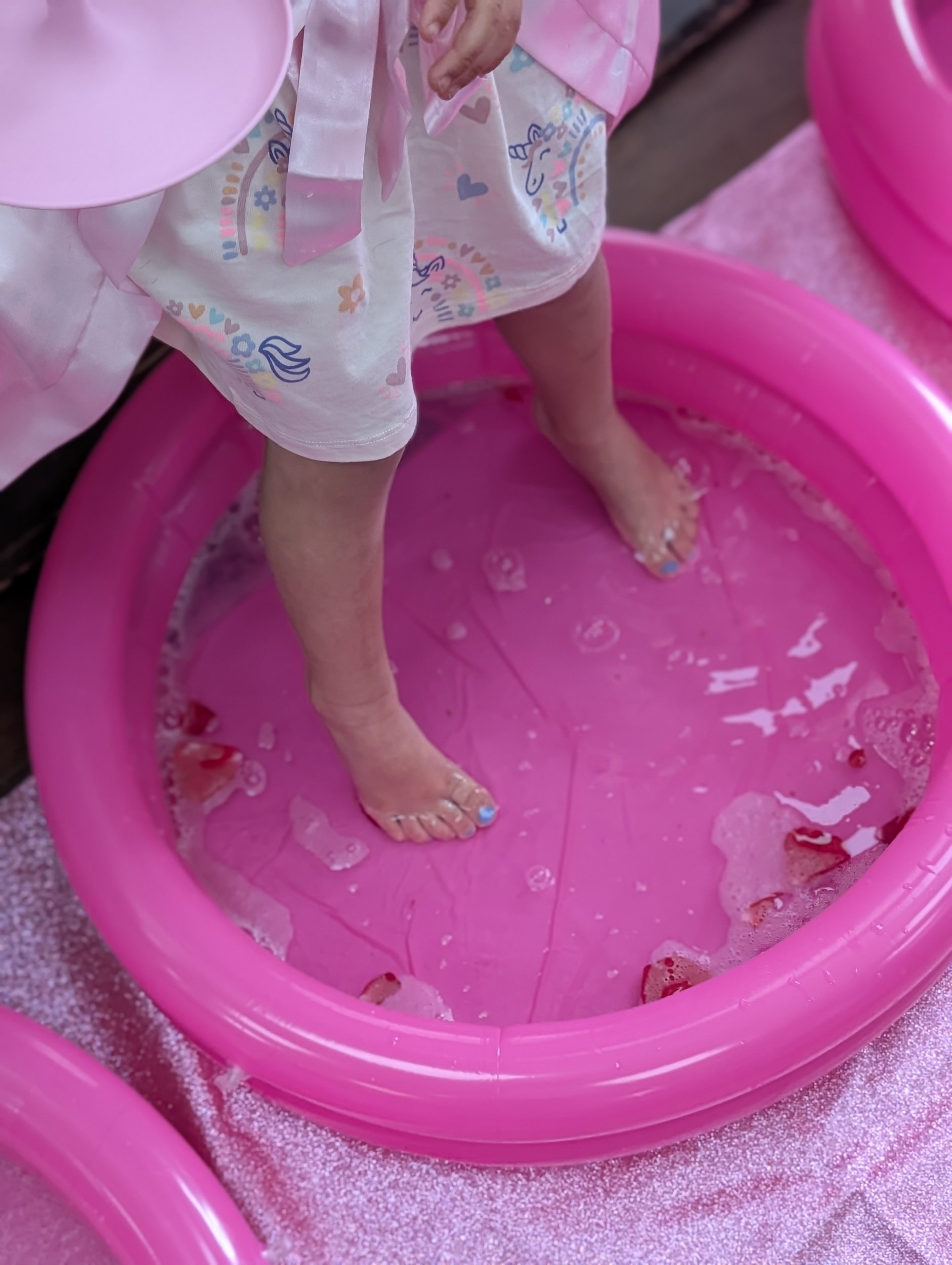 Child standing in a pink plastic inflatable pool with her feet submerged in water, wearing colorful pajamas with unicorn and heart patterns.