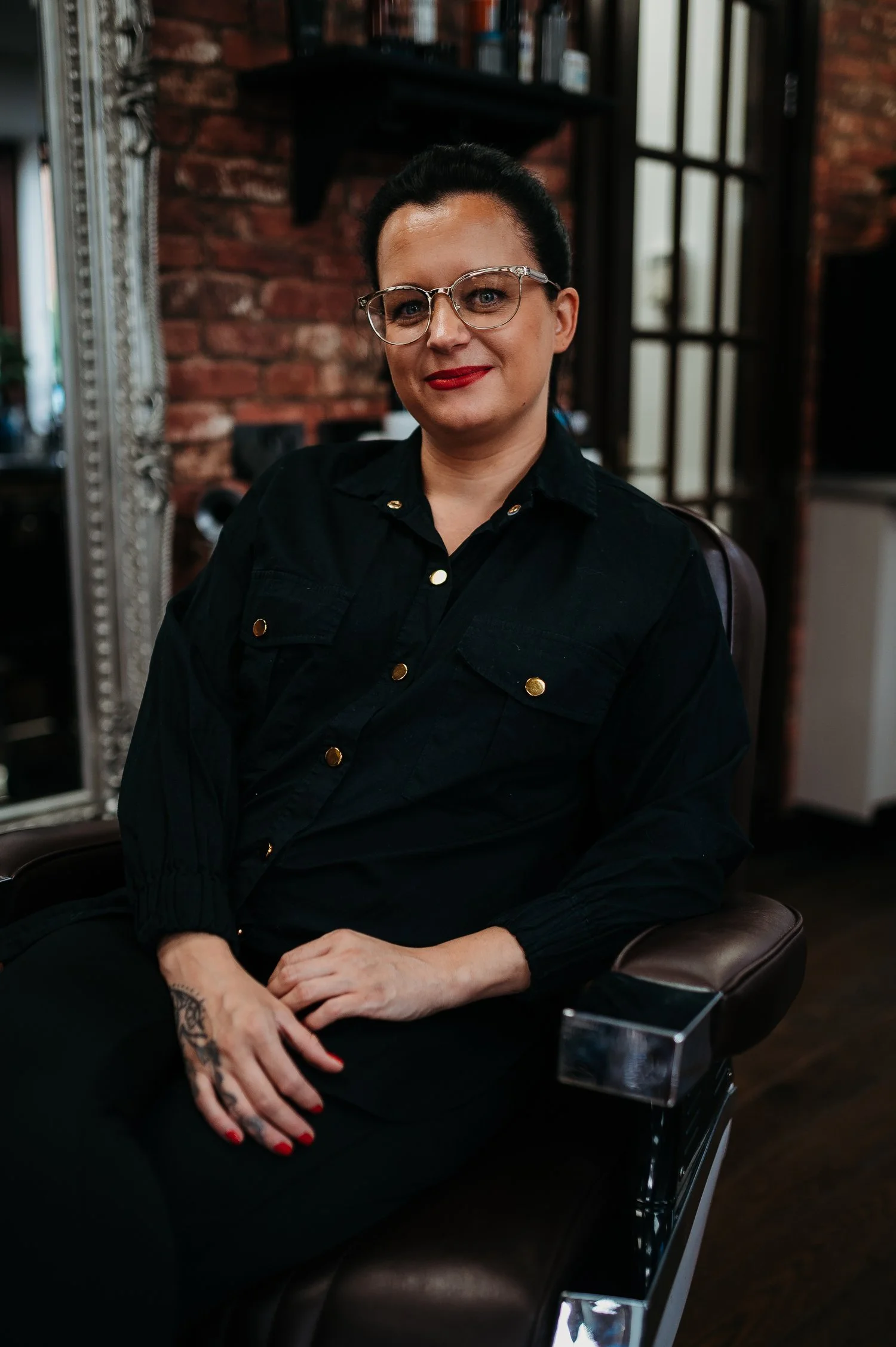 A woman with dark hair, glasses, and red lipstick sitting in a salon chair, wearing a black shirt with gold buttons, with a brick wall and mirror in the background.