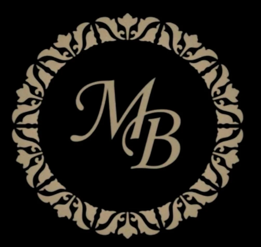 A black circular logo with beige ornate border and the initials 'M B' in the center.