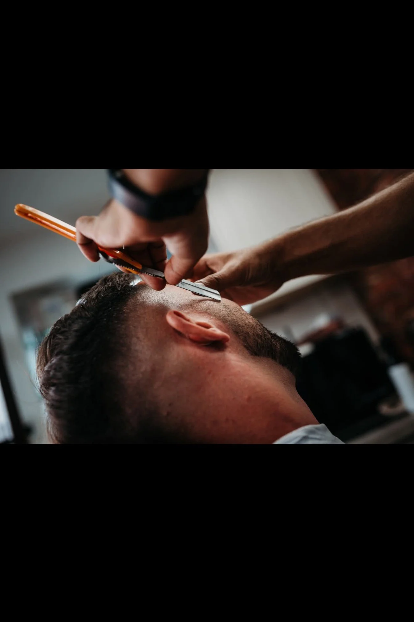 A barber is trimming a man's hair with scissors and clippers in a salon.