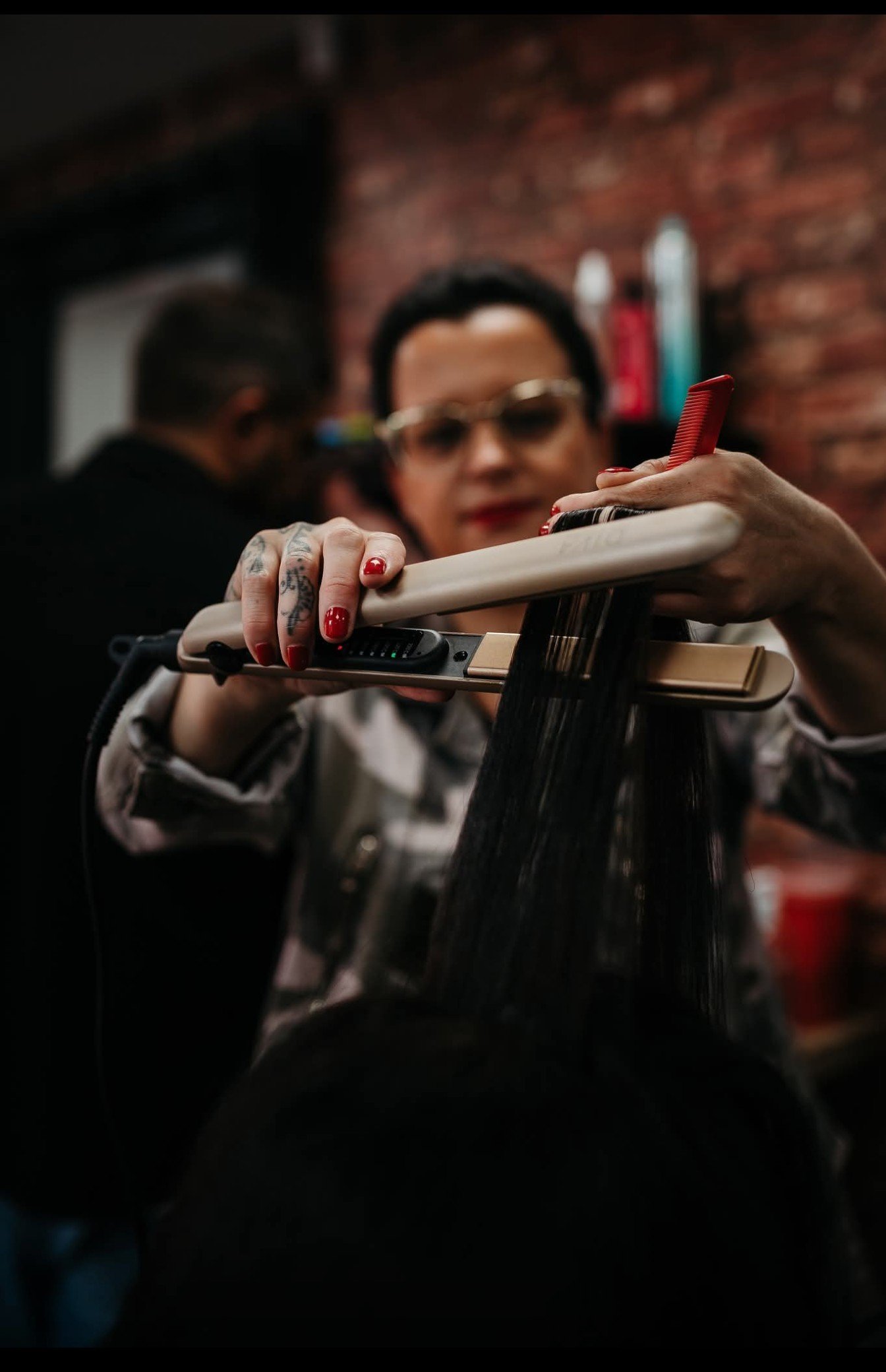 A person with tattoos on their fingers and red-painted nails using a flat iron to straighten black hair in a salon with a brick wall in the background.