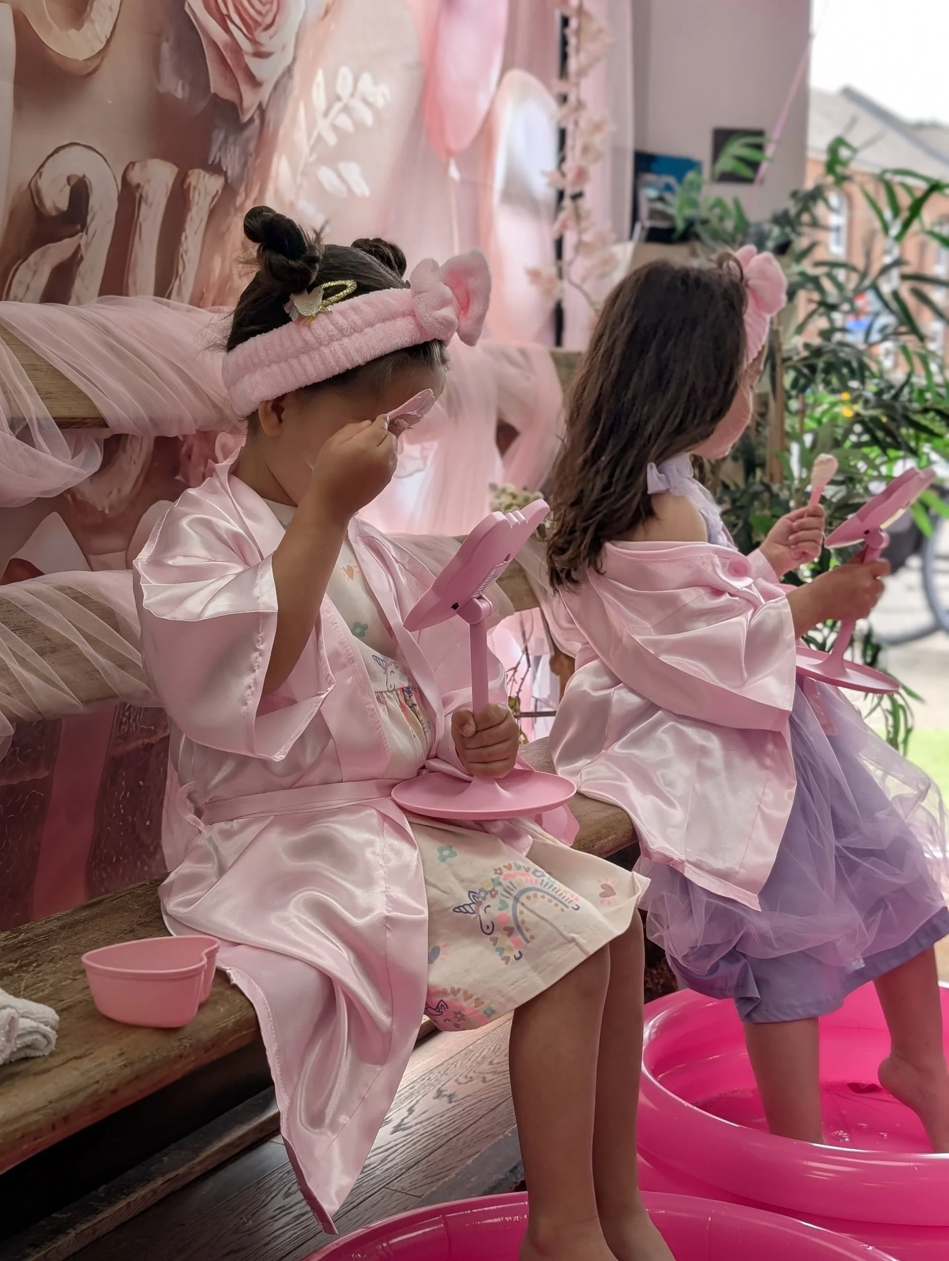 Two young girls in pink robes and headbands with ears are sitting on a wooden bench, each holding a small pink mirror. One girl is wiping her face with a cloth, and the other is looking into her mirror. There is a large pink basin on the floor filled with water in front of them, and a pink bowl on the bench. The background shows a decorated wall with pink elements and some plants near a window.